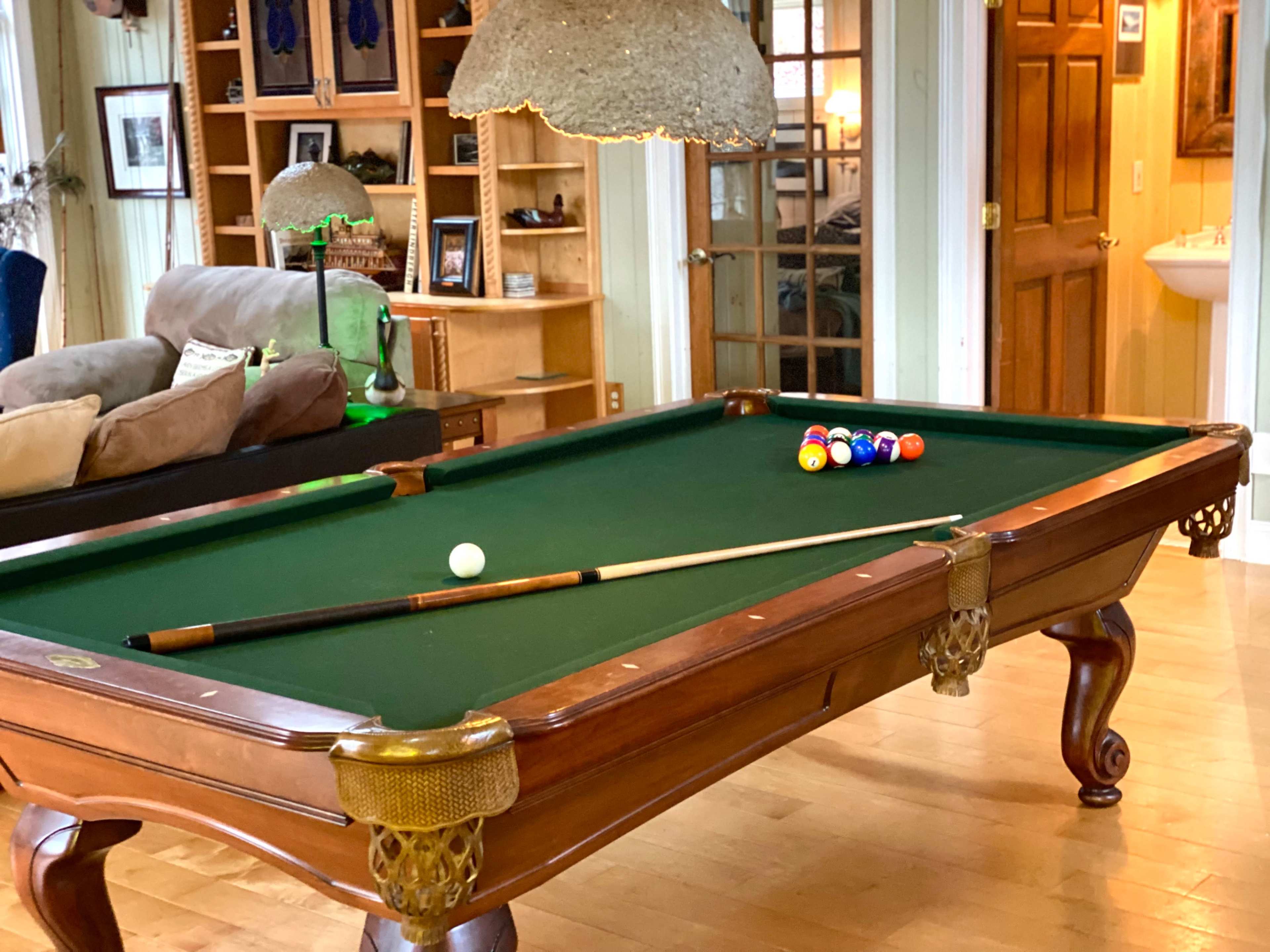 A green-felt billiard table with a cue stick and a set of colored balls arranged on its surface, located in a well-furnished room.
