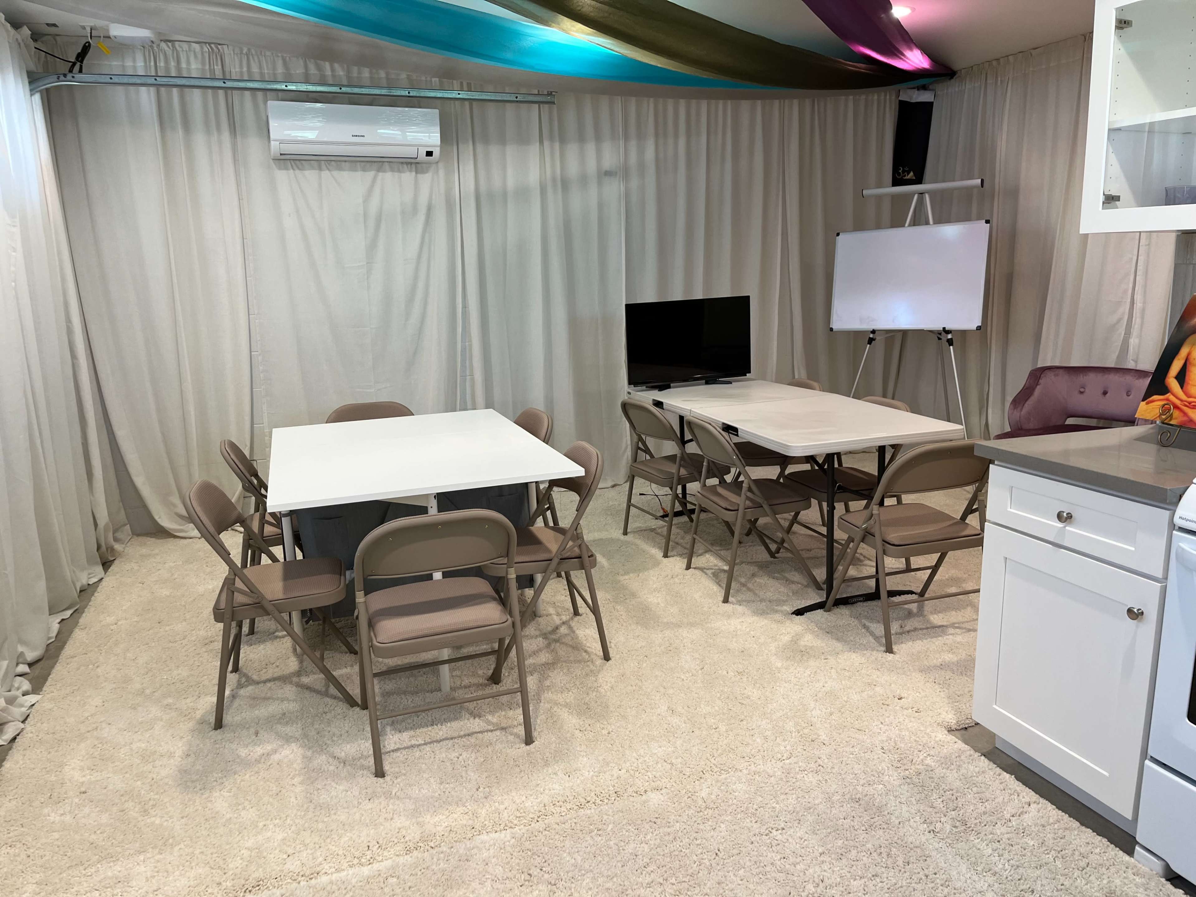 A simple, sparsely furnished room features two folding tables surrounded by chairs, a television, and a whiteboard, all set against draped curtains.