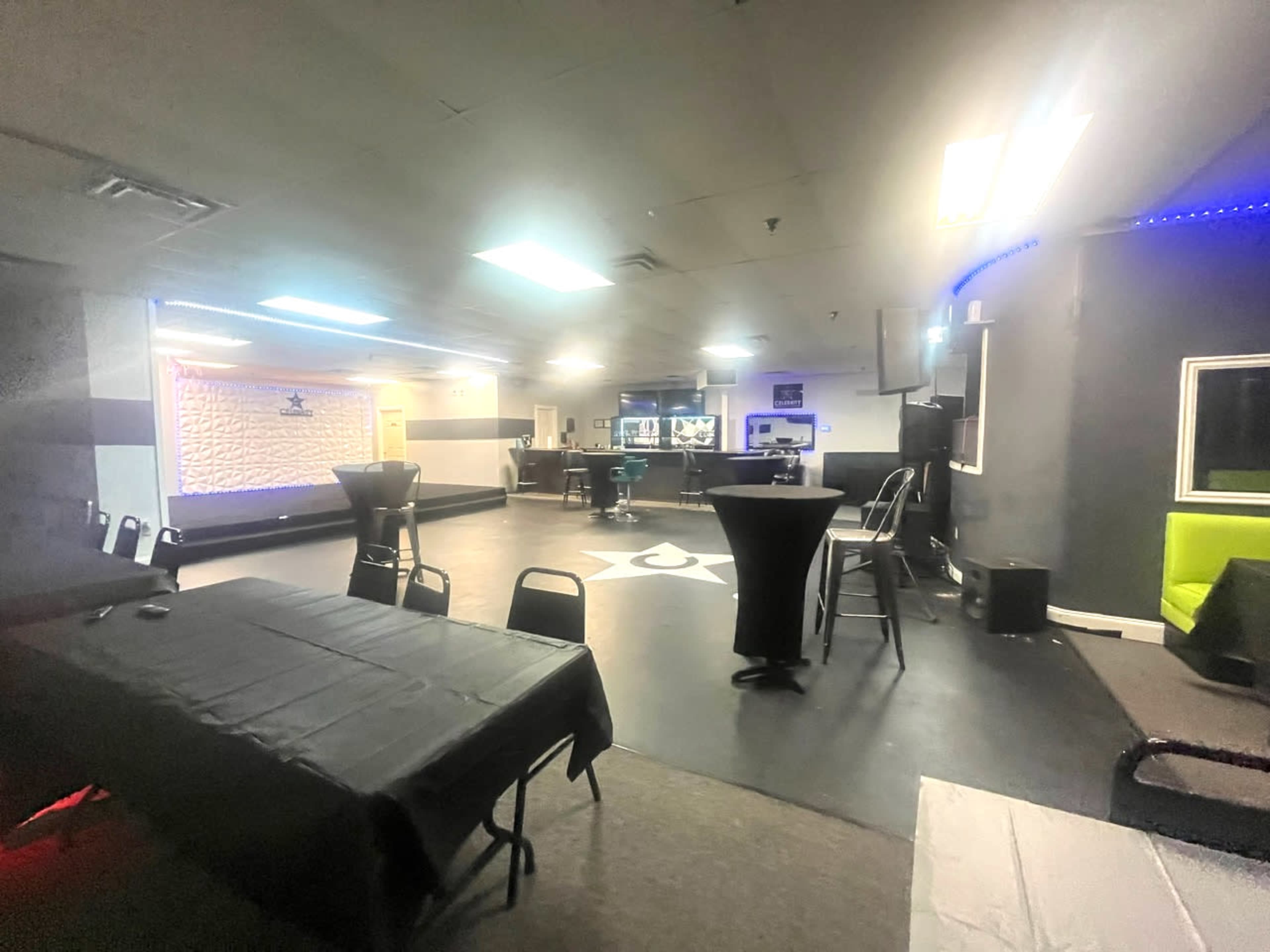 An empty, dimly lit event space with tables and chairs arranged around a central dance floor and a bar at one end.