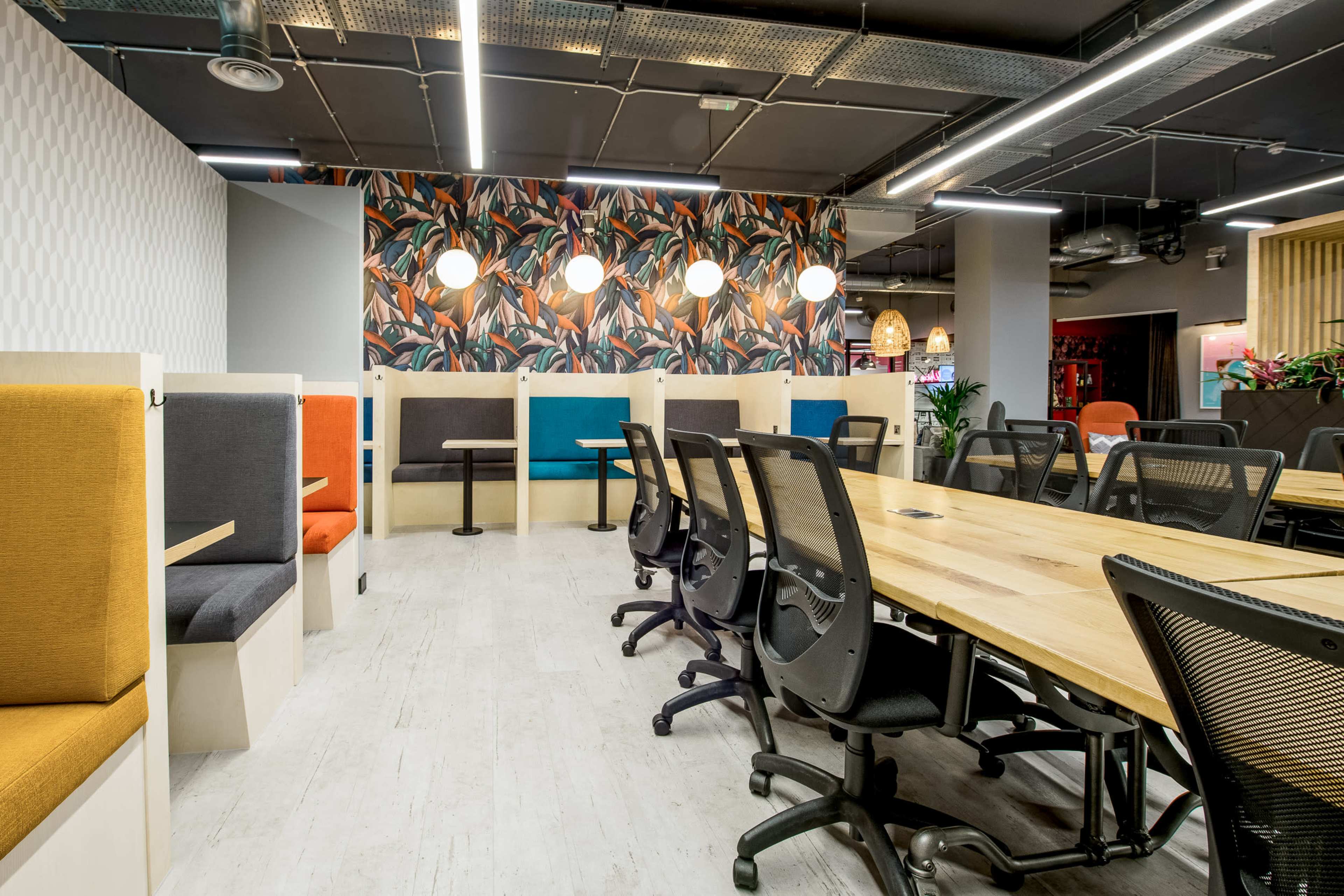 A modern office space with colorful seating areas, large wooden tables, and decorative wallpaper featuring tropical leaves.