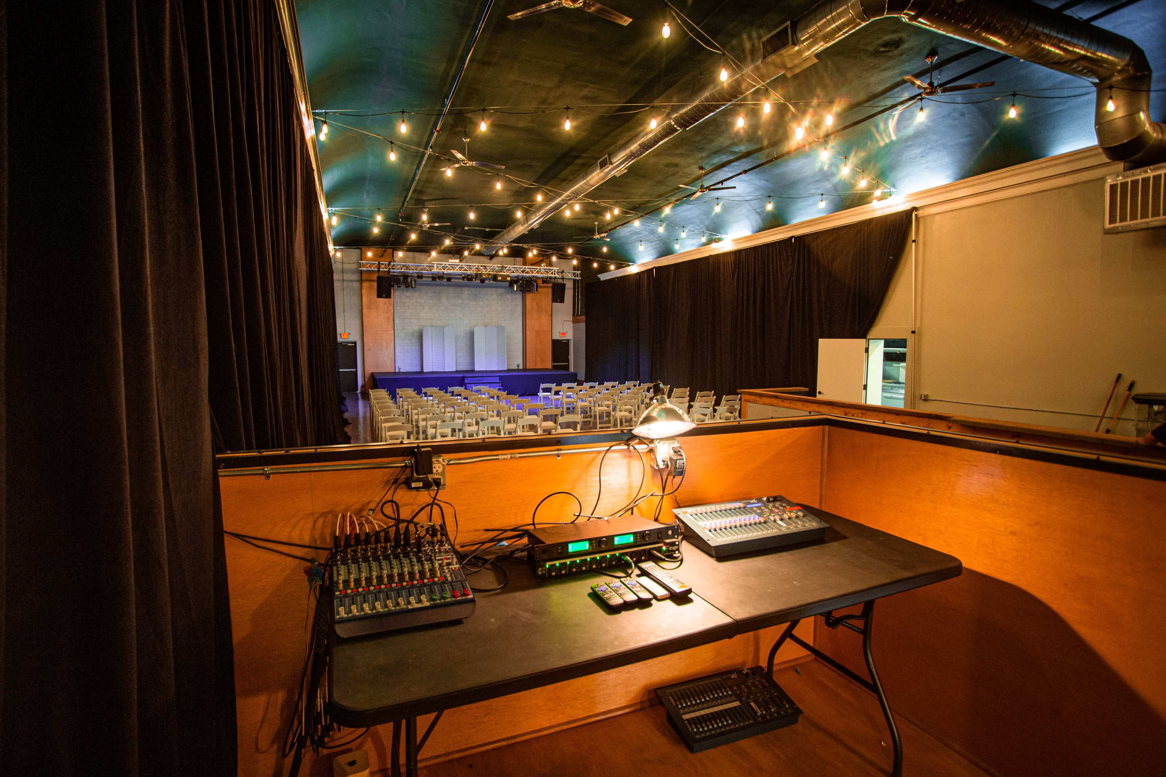 The image shows a sound mixing station in a dimly lit theater, with chairs arranged for an audience in the background and stage lights hanging above.