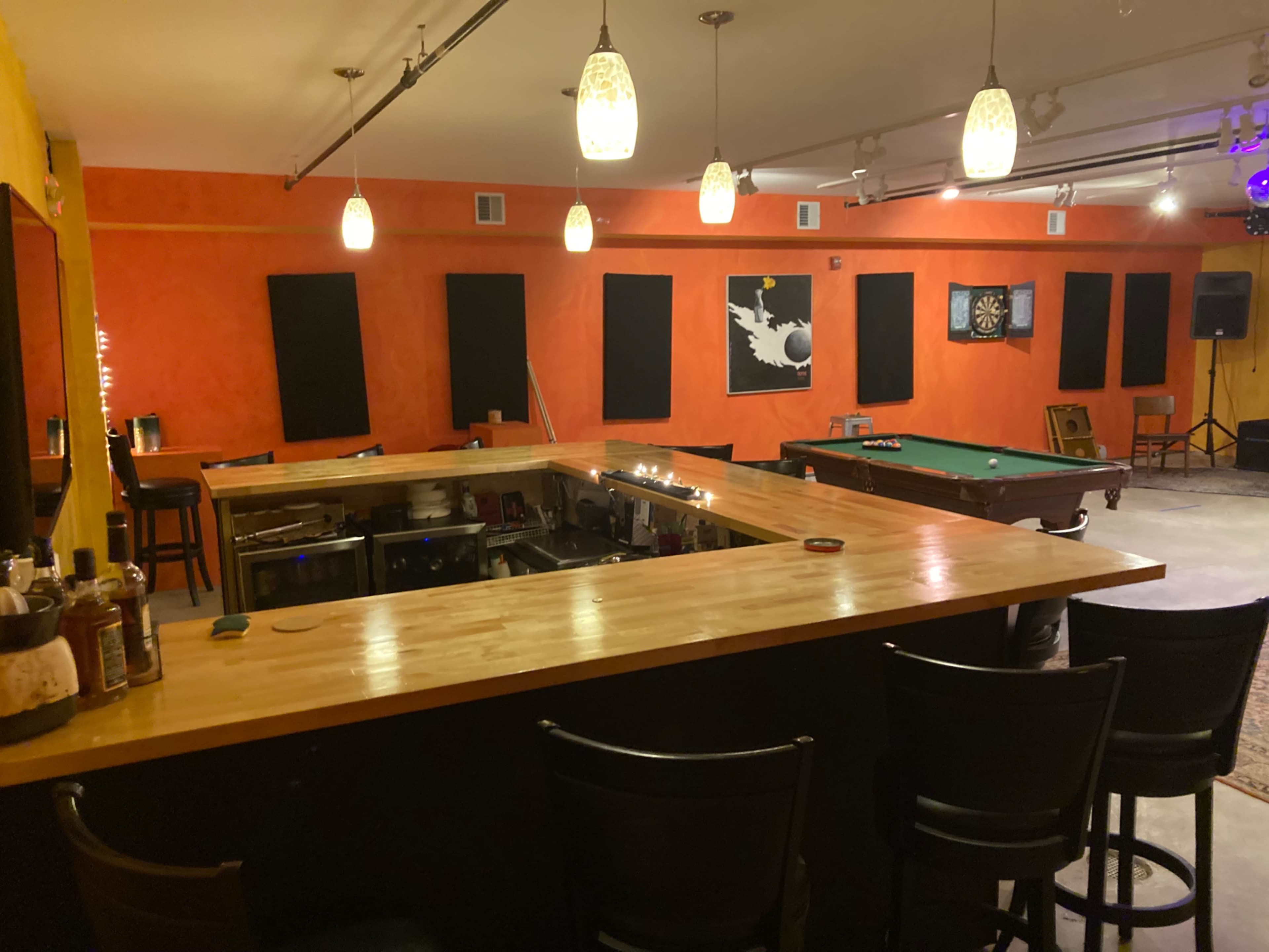 A bar area with a wooden counter, several black chairs, a pool table, and orange walls adorned with black window coverings and a piece of artwork.