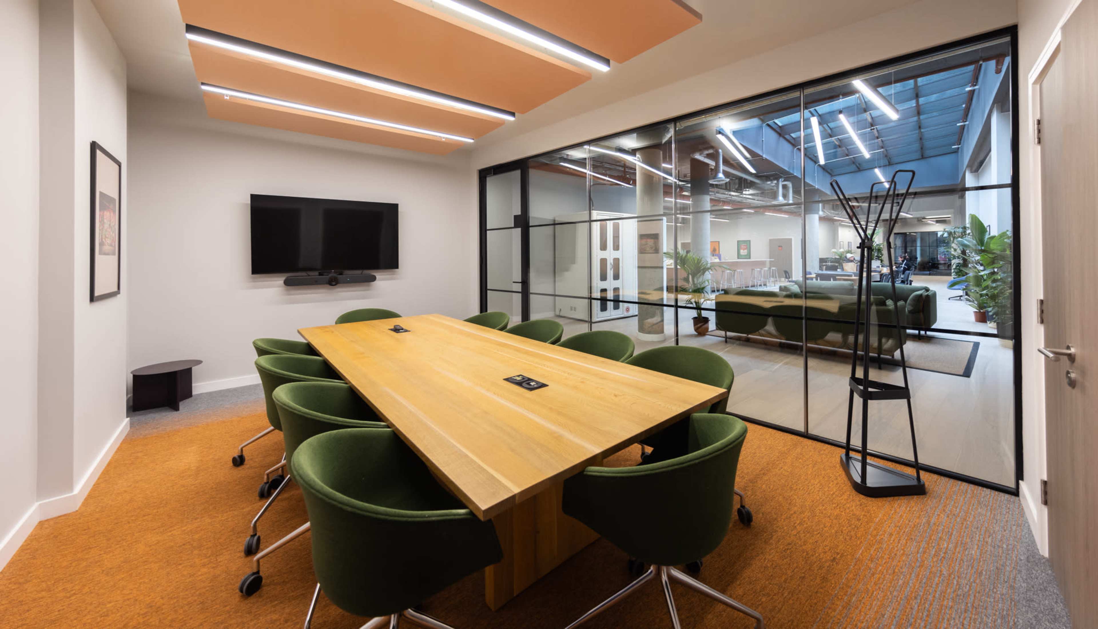 A modern conference room features a long wooden table surrounded by green chairs, with large glass walls revealing an open office space.