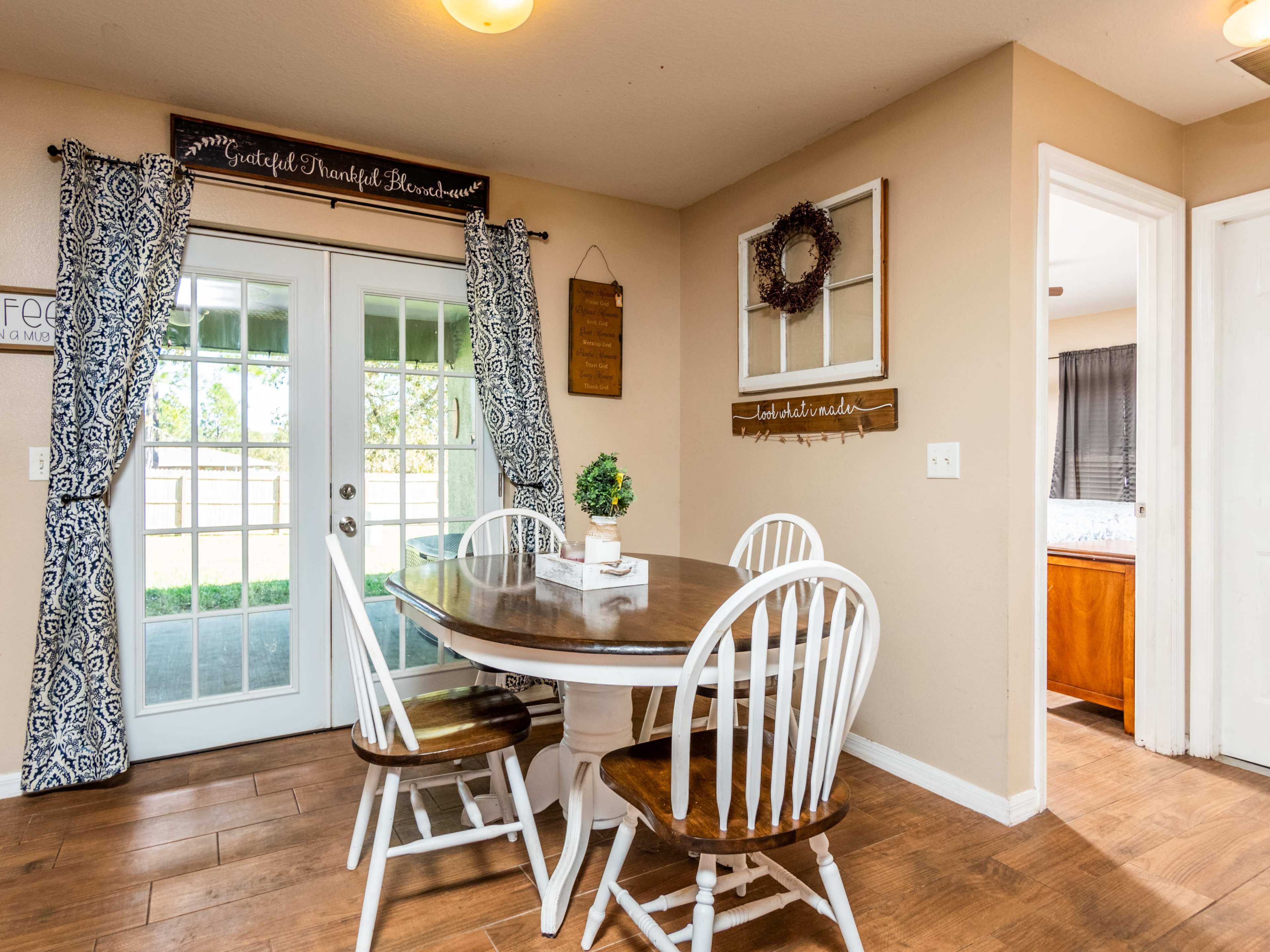 A dining area features a round wooden table with four chairs, surrounded by beige walls and large windows with patterned curtains.