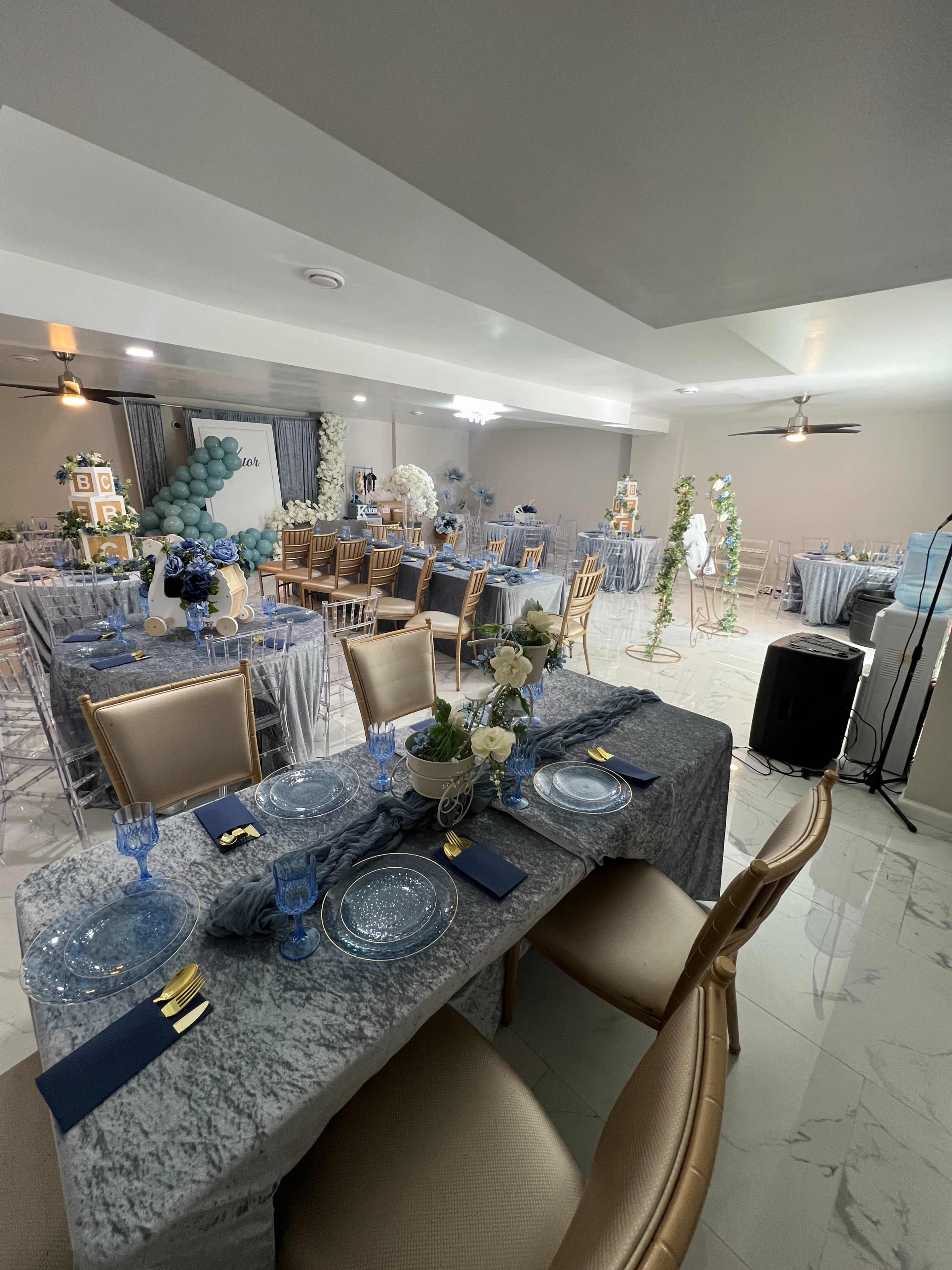 The image shows a beautifully arranged event space featuring tables set with blue and gold tableware, decorated with flowers and balloons, and illuminated by soft lighting.