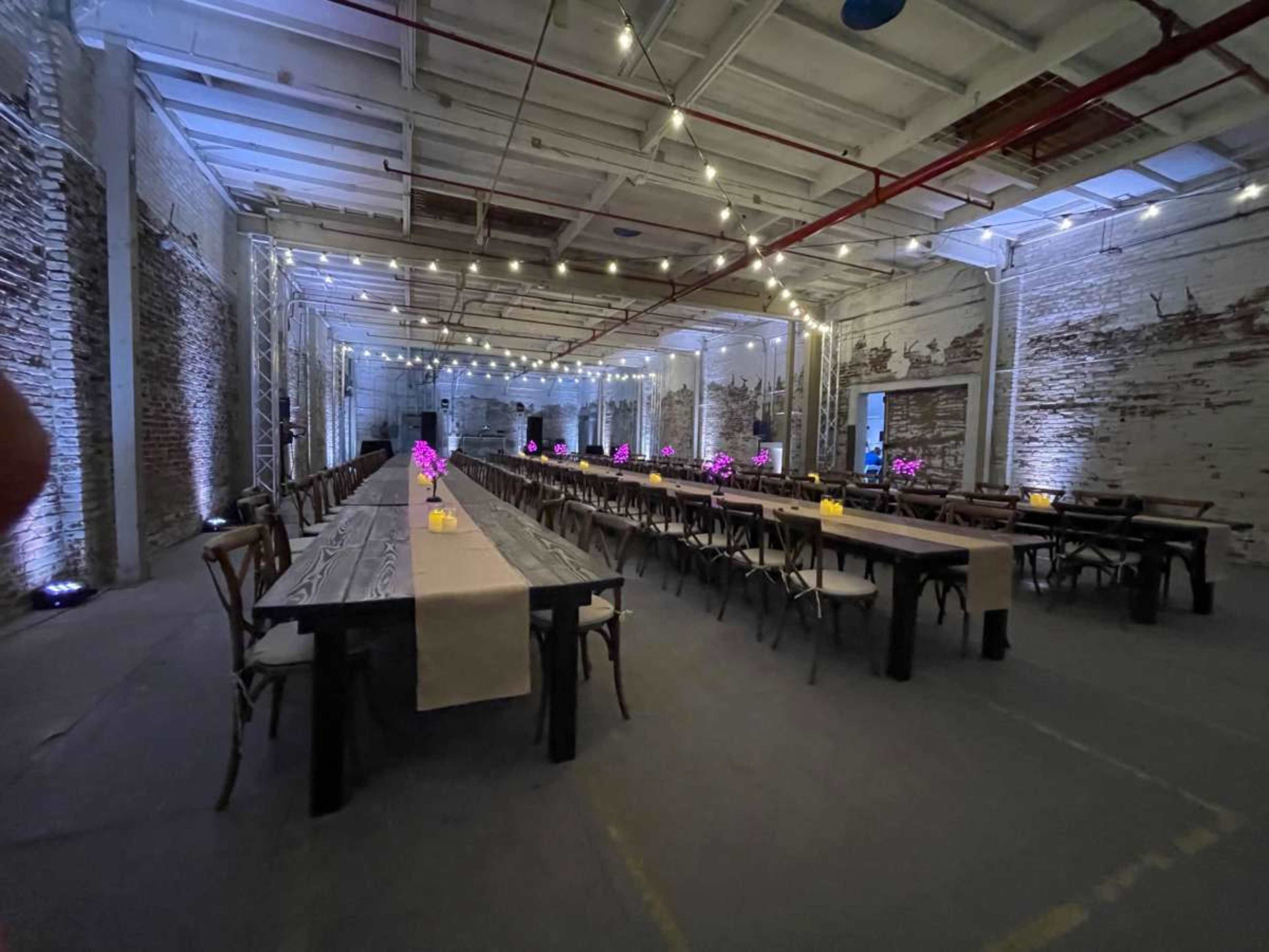 The image shows a spacious industrial hall set up for an event, featuring long tables with floral centerpieces and soft lighting.