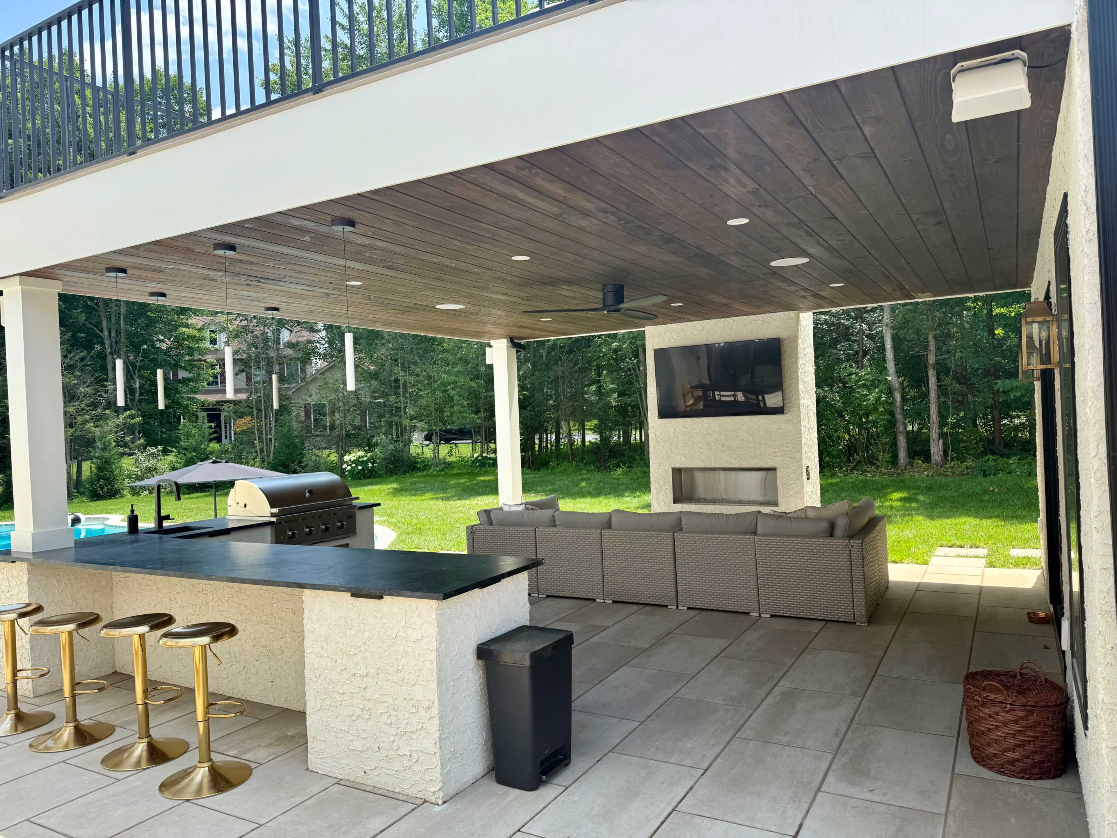The image shows an outdoor patio featuring a kitchen area with a grill, a seating arrangement, and a television under a wooden ceiling, surrounded by lush greenery.