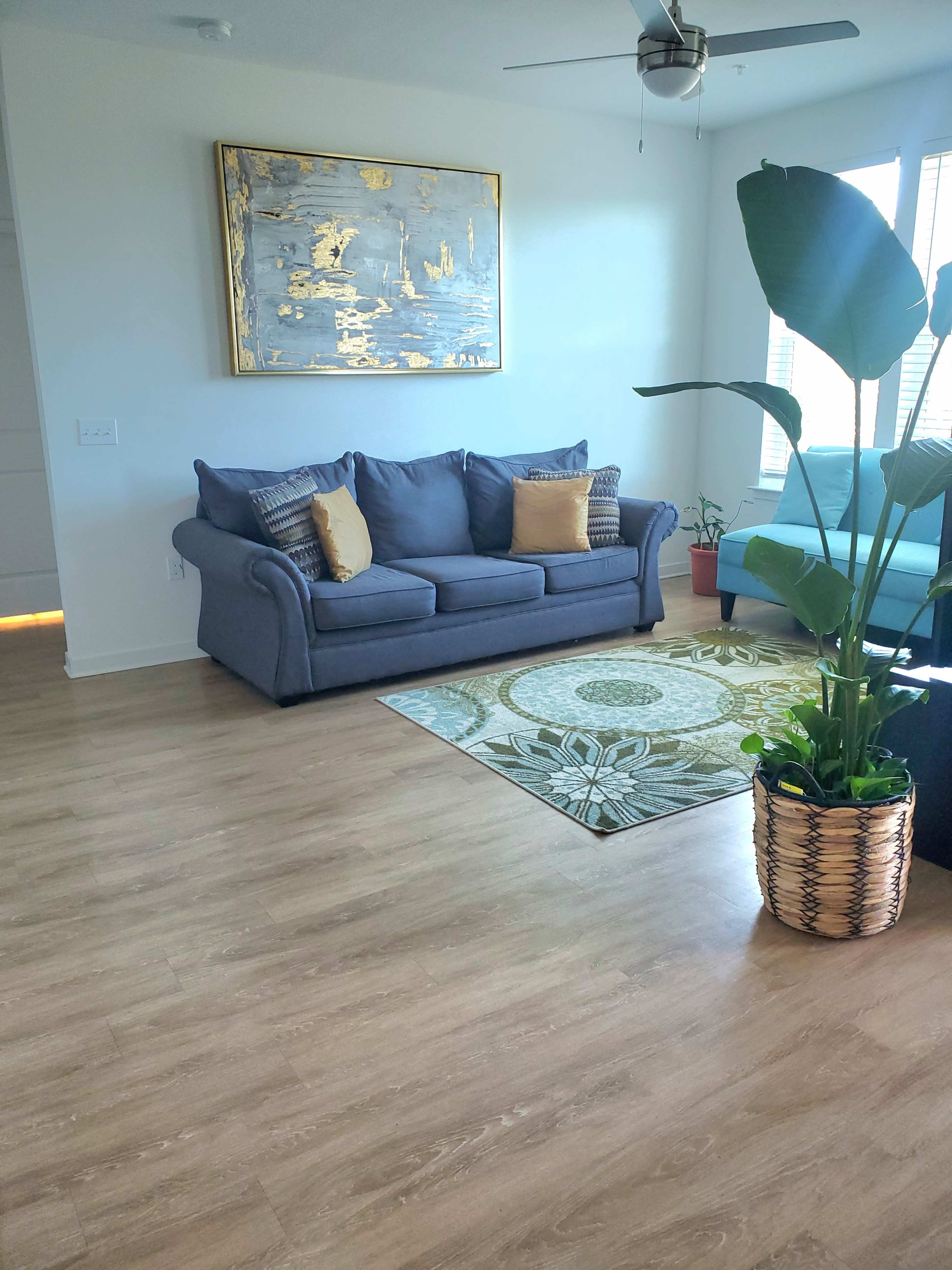 A living room with a blue sofa, a green armchair, a patterned area rug, a large decorative wall art piece, and a potted plant.