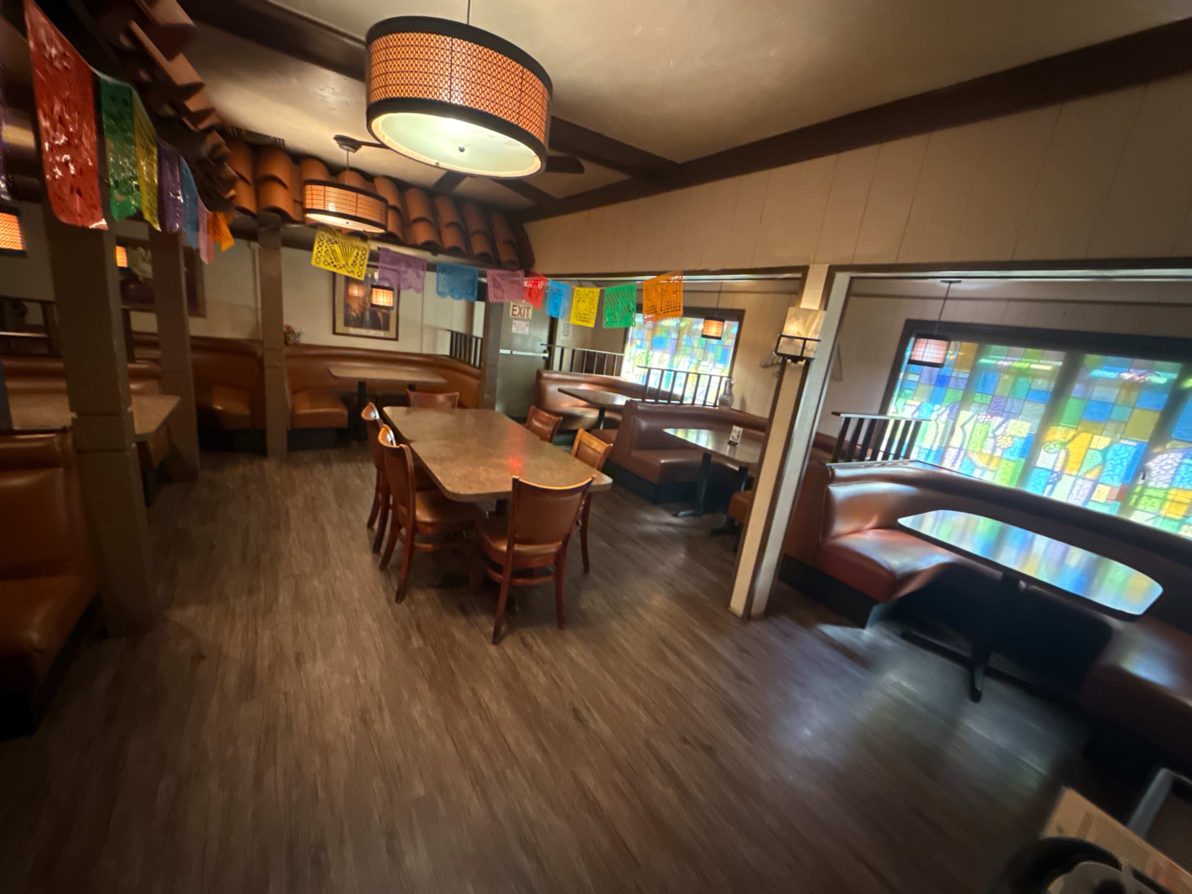 The image shows a spacious restaurant interior featuring wooden booths and tables, adorned with colorful papel picado banners and stained glass windows.
