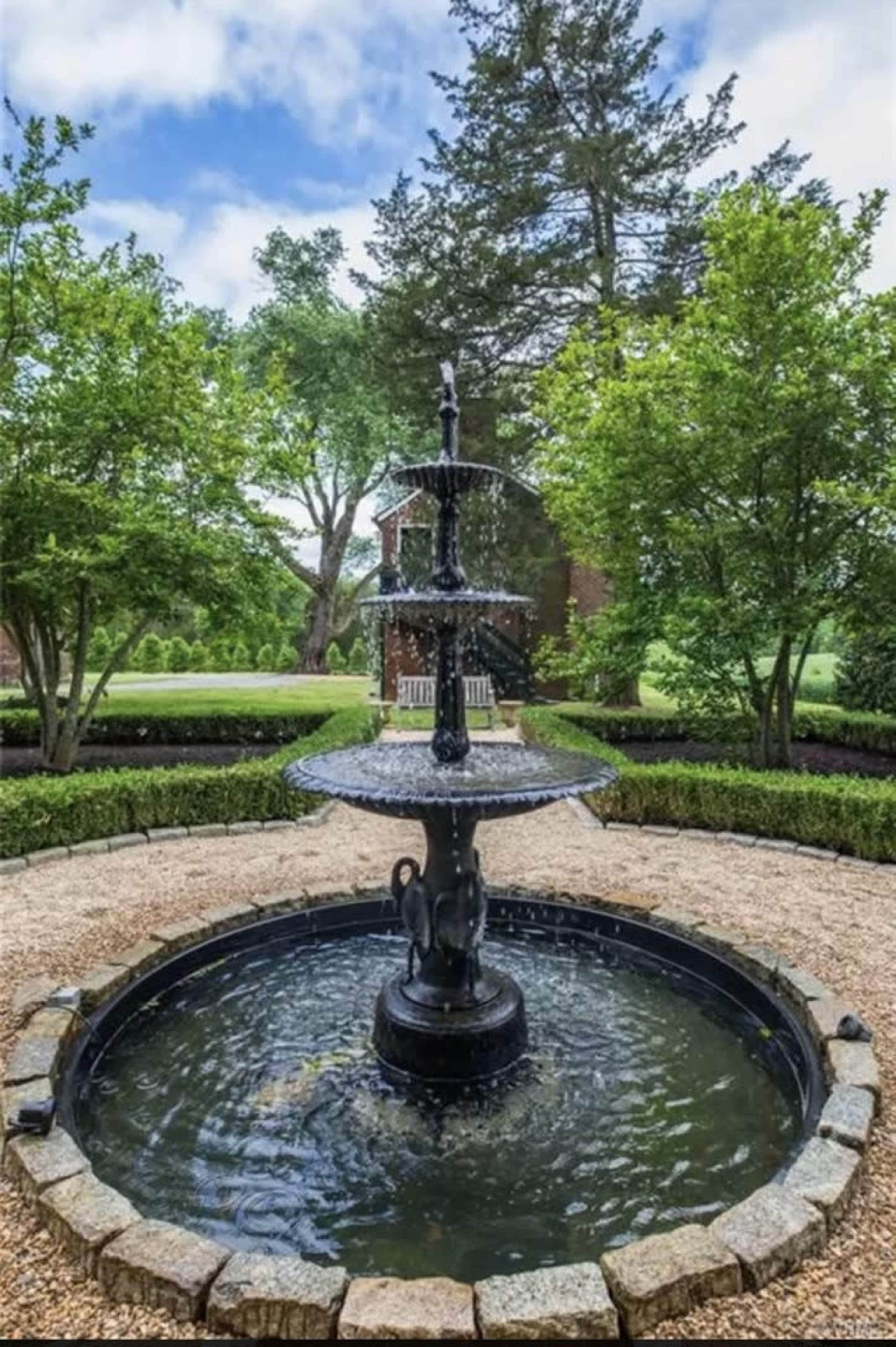 A multi-tiered fountain is situated in a circular stone basin surrounded by lush greenery and a well-maintained garden.