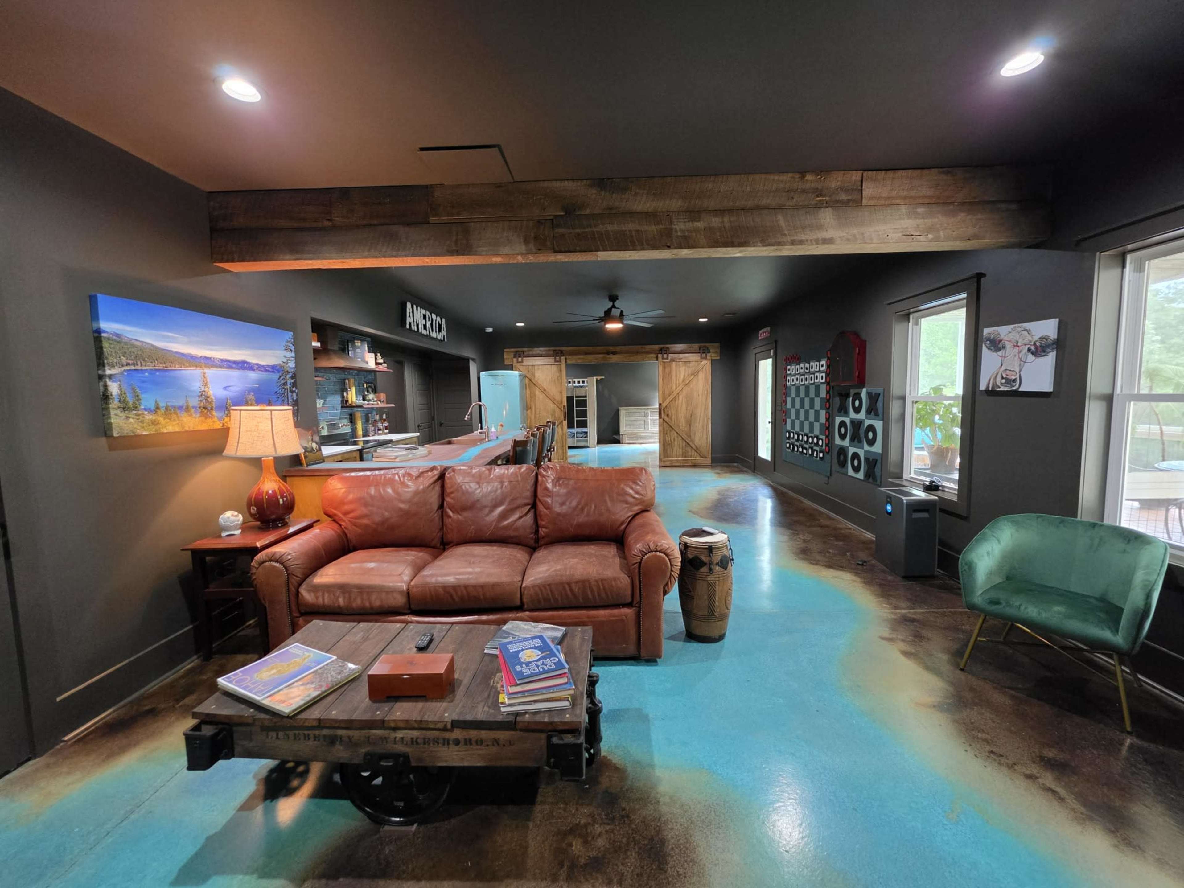 The image shows a cozy living room area with a brown leather couch, a coffee table, and a vibrant blue floor, complemented by a kitchenette and large windows.