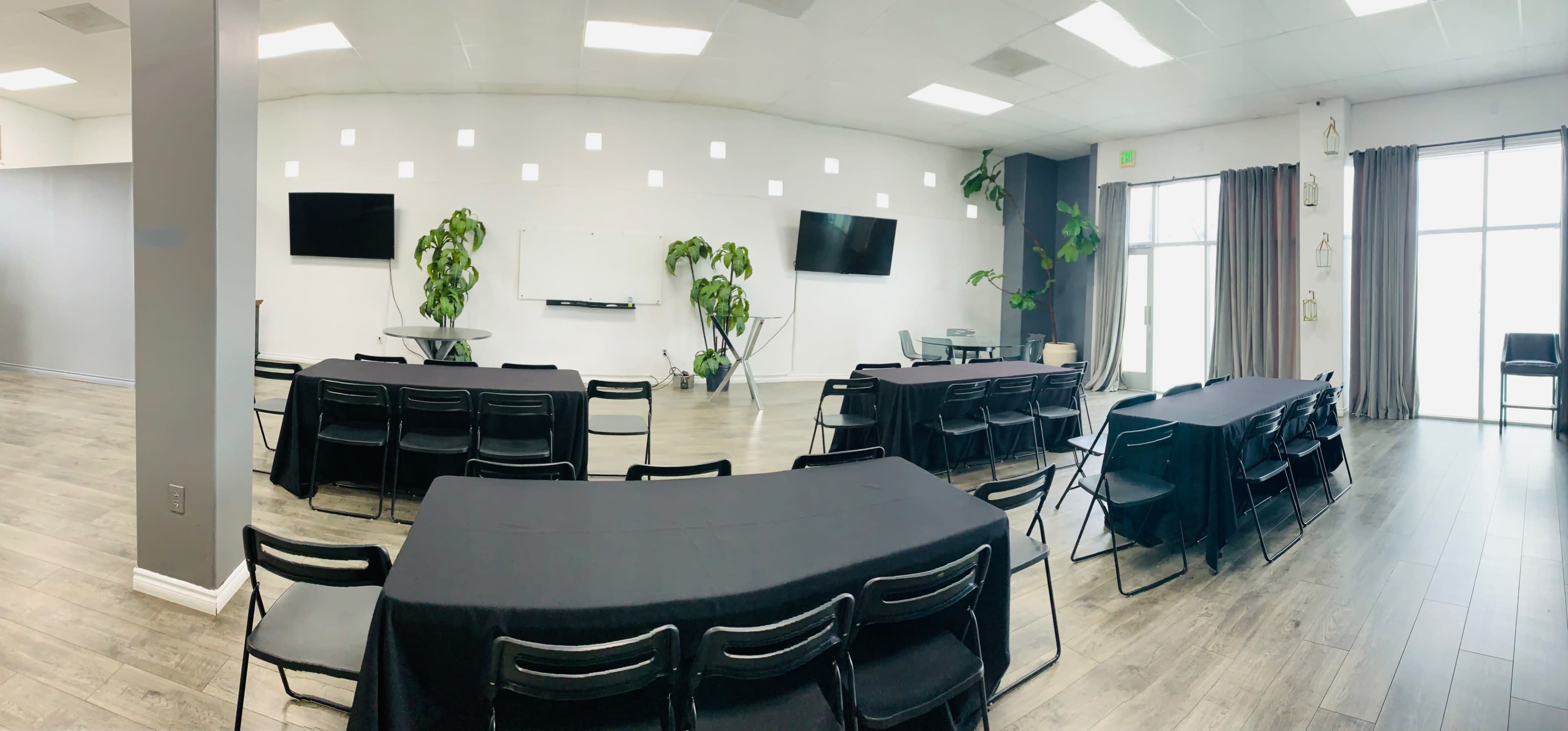 A spacious meeting room arranged with several tables and chairs, flanked by large windows and equipped with wall-mounted TVs and indoor plants.