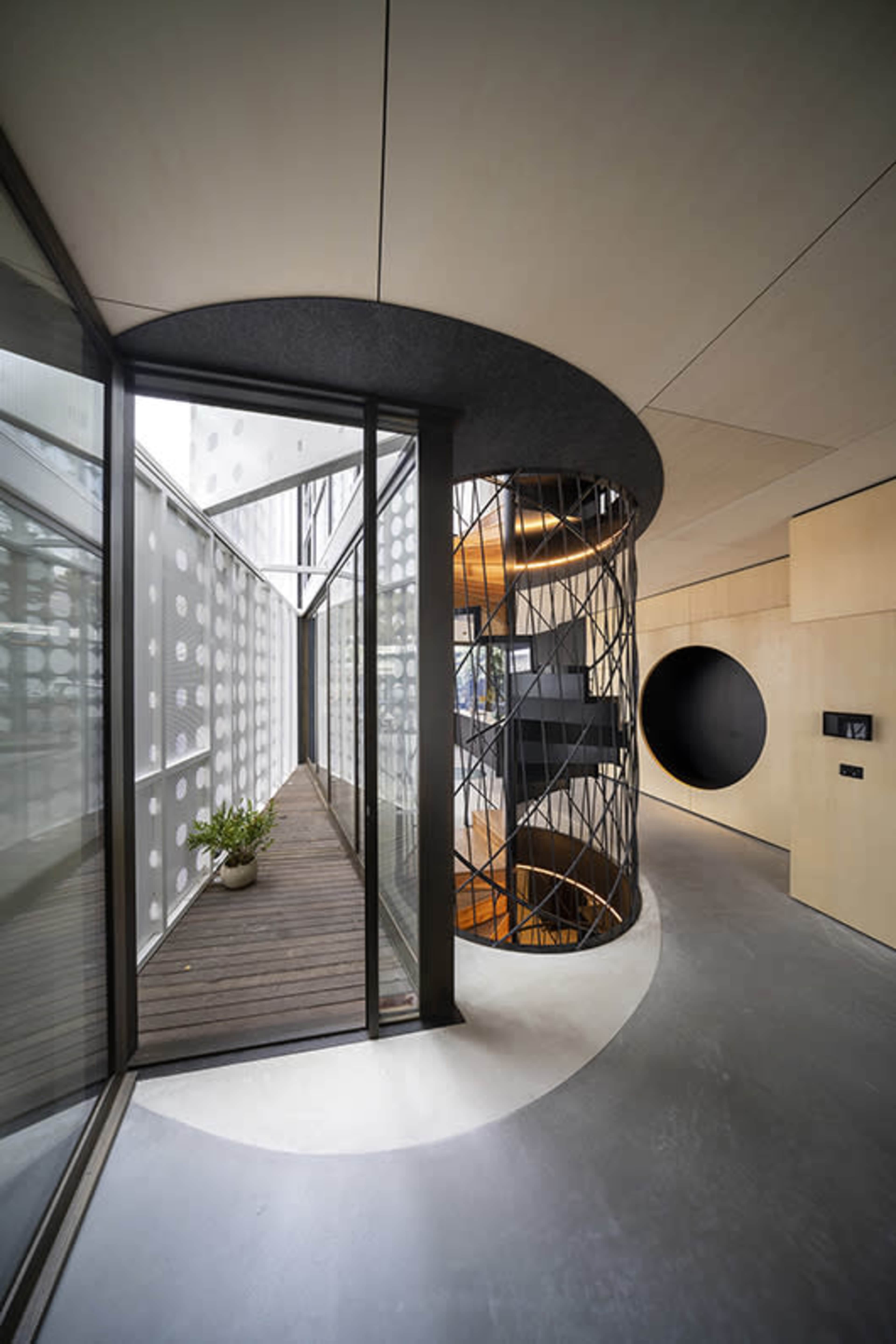 A modern interior hallway featuring a circular staircase, a wall with circular cutouts, and large glass panels allowing natural light.