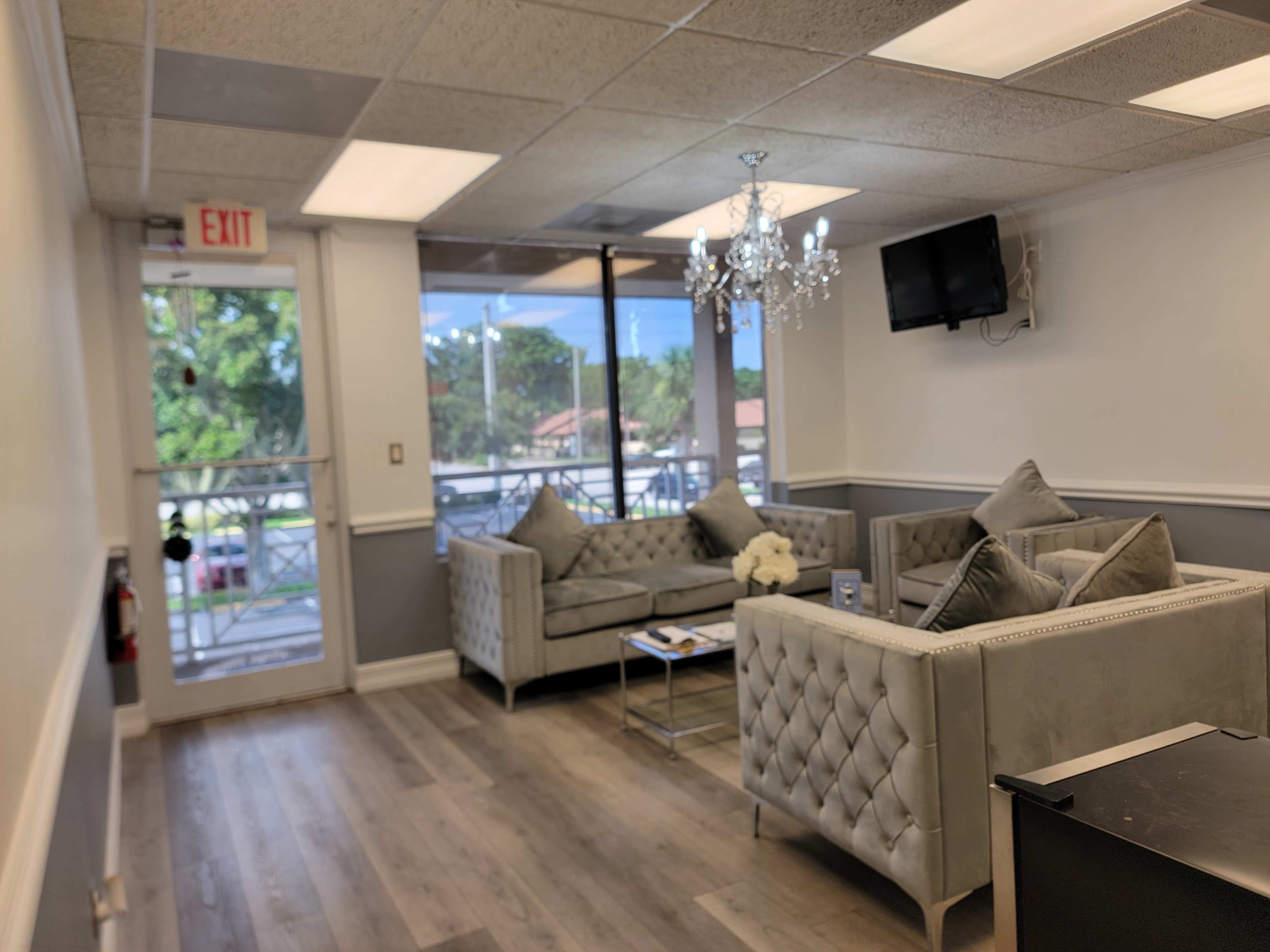 The image shows a waiting area with two gray tufted couches, a chandelier, a television, and large windows letting in natural light.