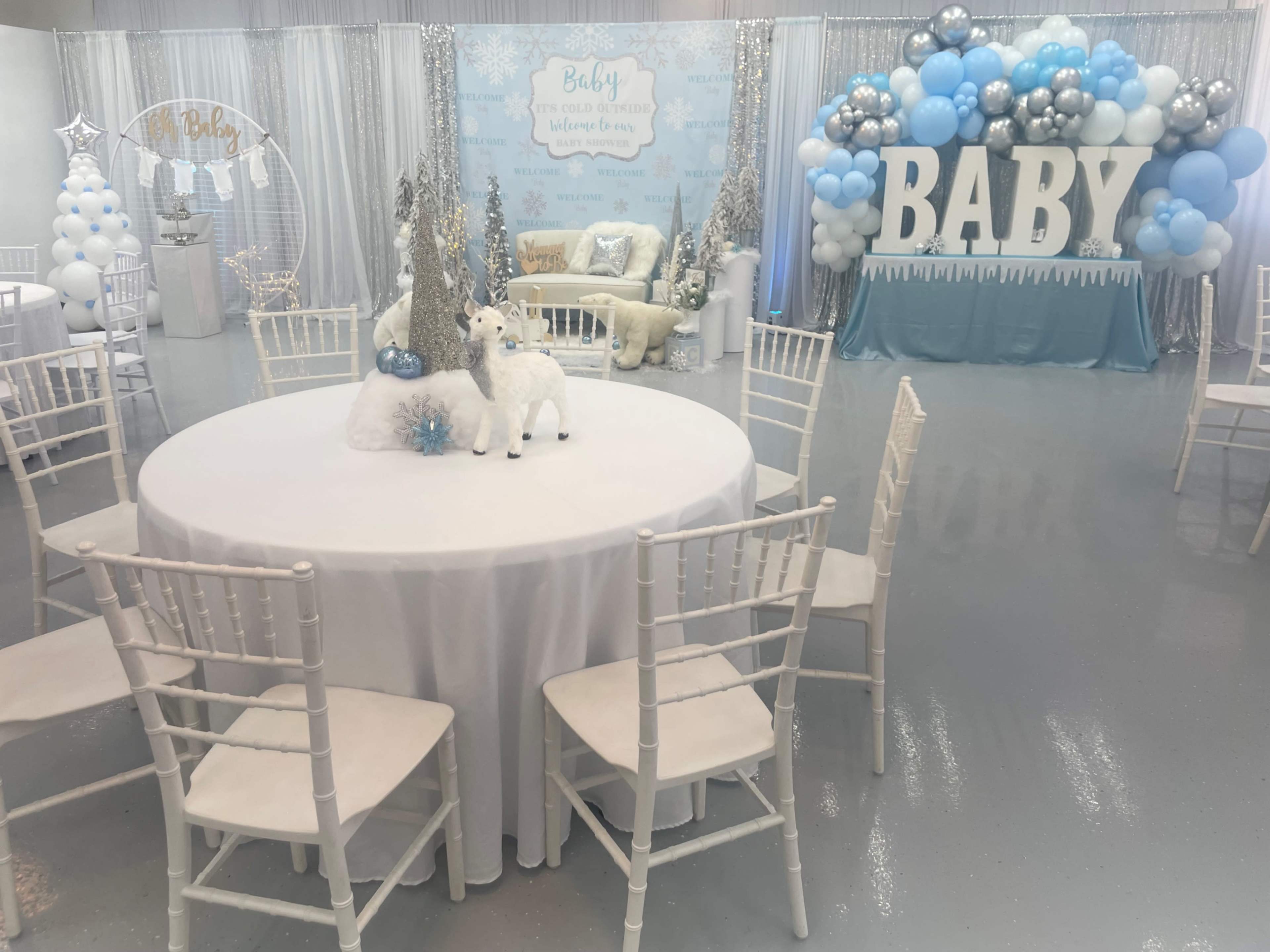 A decorated event space for a baby shower features a round table with a white tablecloth and centerpieces, surrounded by white chairs, and adorned with snow-themed decorations and a balloon arch displaying the word "BABY."