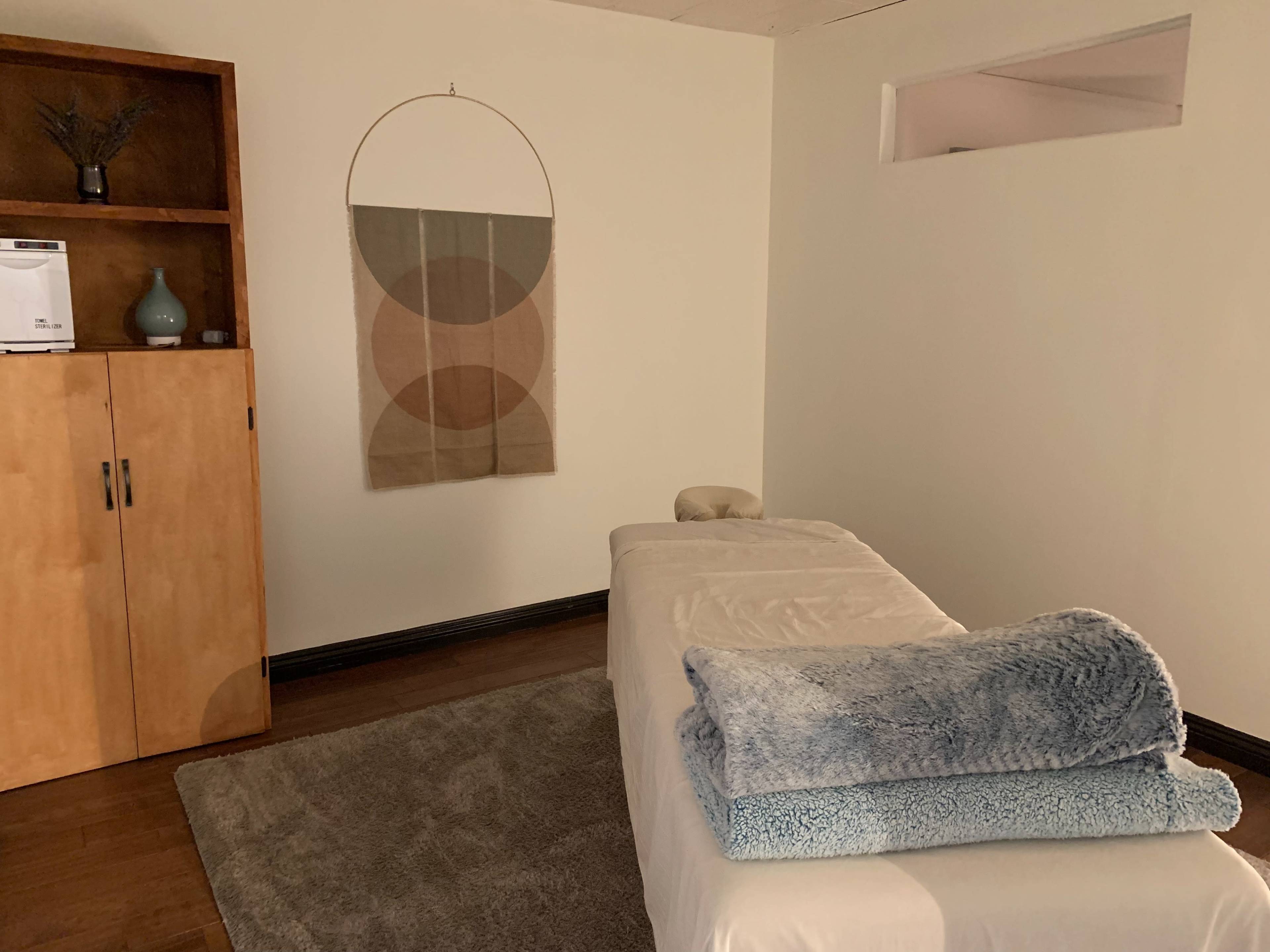 The image shows a massage therapy room featuring a treatment table topped with a folded blanket, a wooden cabinet, and a wall hanging.