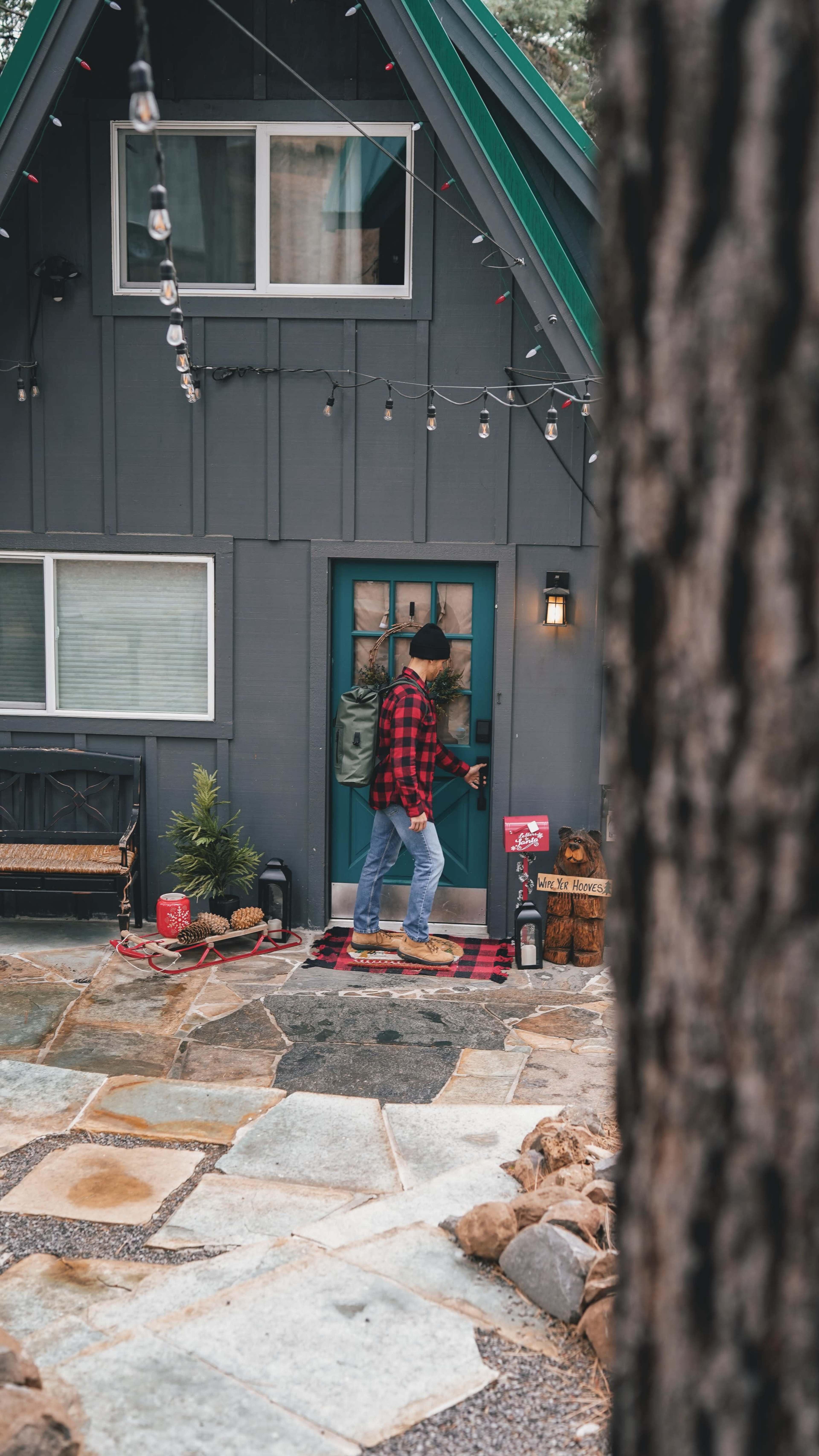 A person wearing a red flannel shirt walks towards the door of a gray cabin with a green roof in a wooded area.