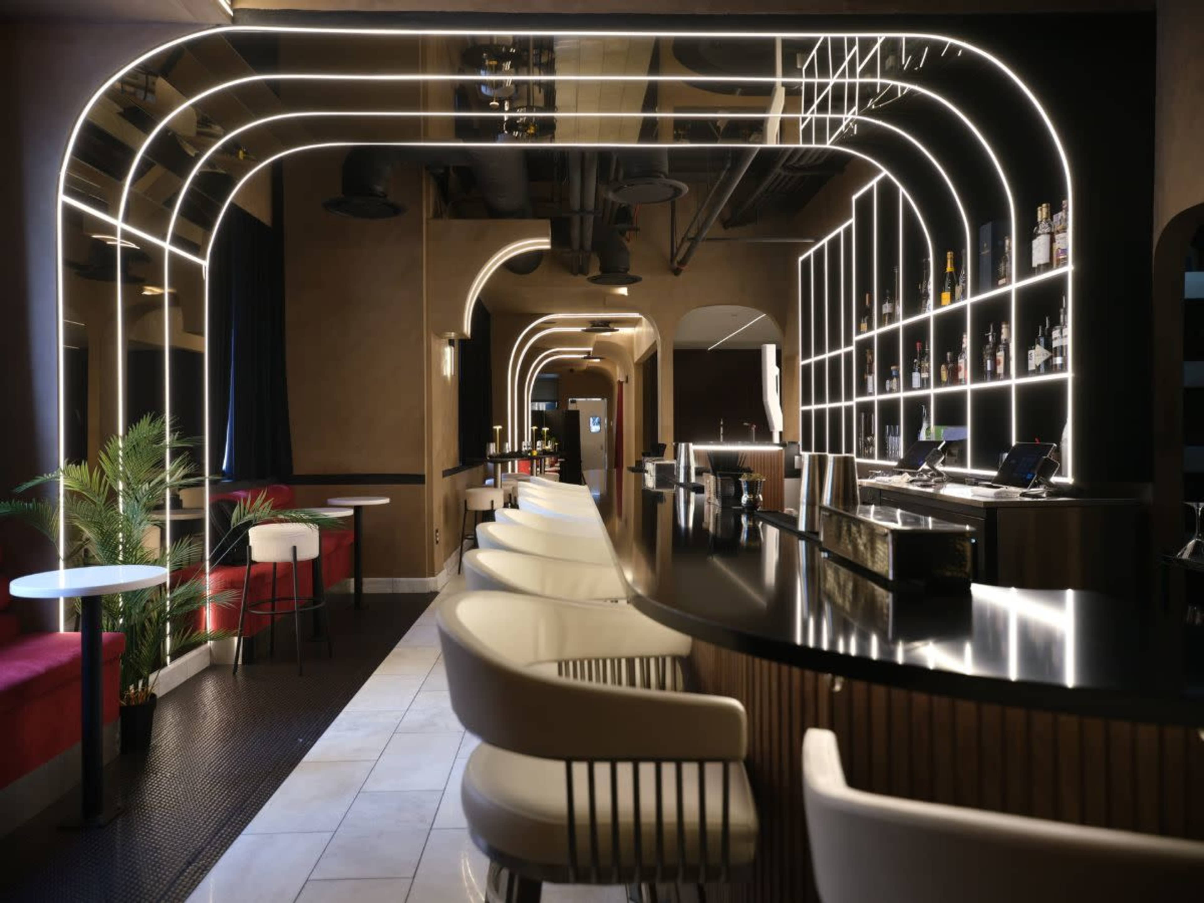 The image shows a modern bar interior with curved lighting, a long counter with white stools, and shelves displaying various bottles in the background.