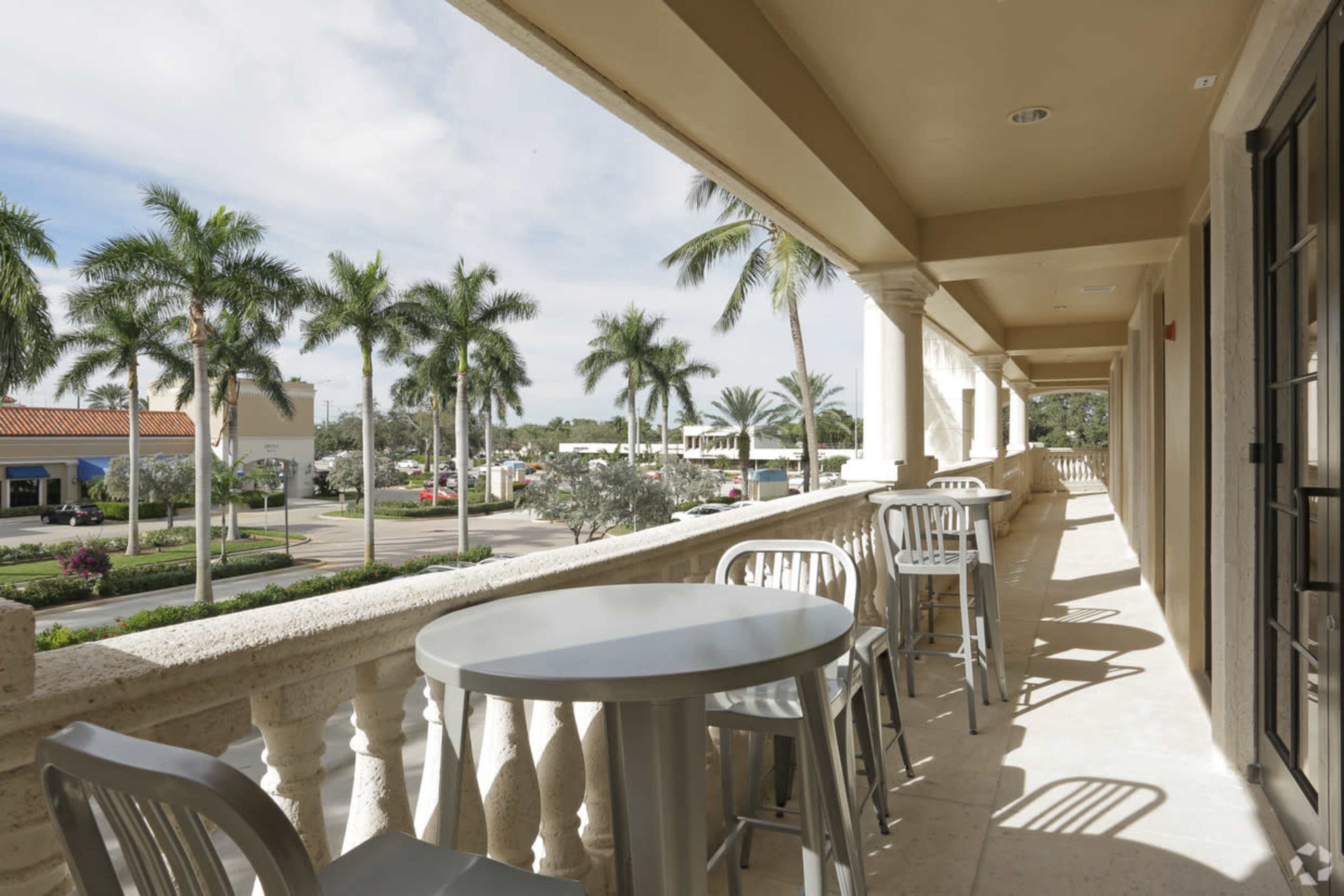 The image shows a balcony with grey tables and chairs overlooking a palm tree-lined street.