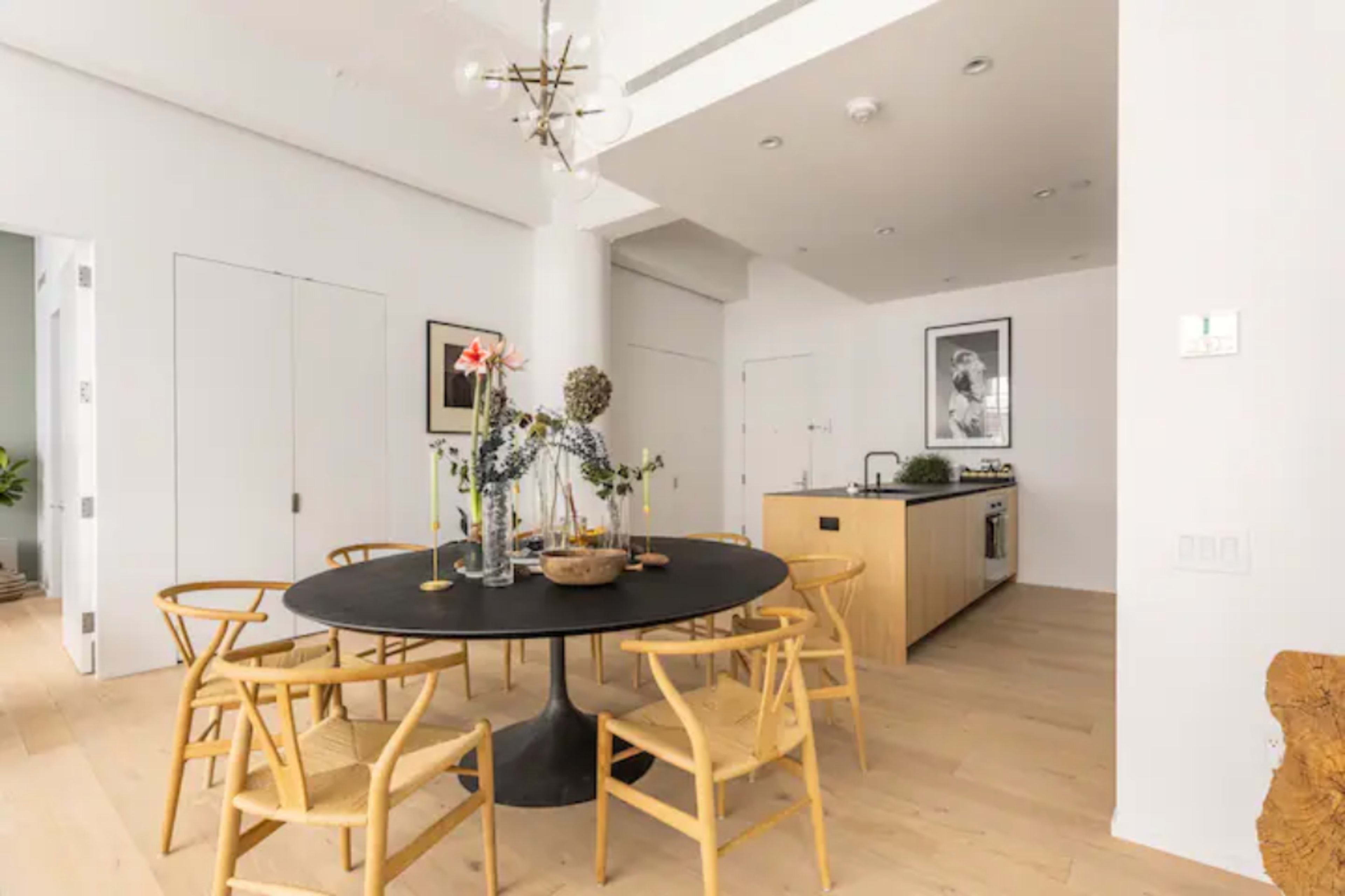 A modern dining area features a round black table surrounded by wooden chairs, with a small kitchen in the background.
