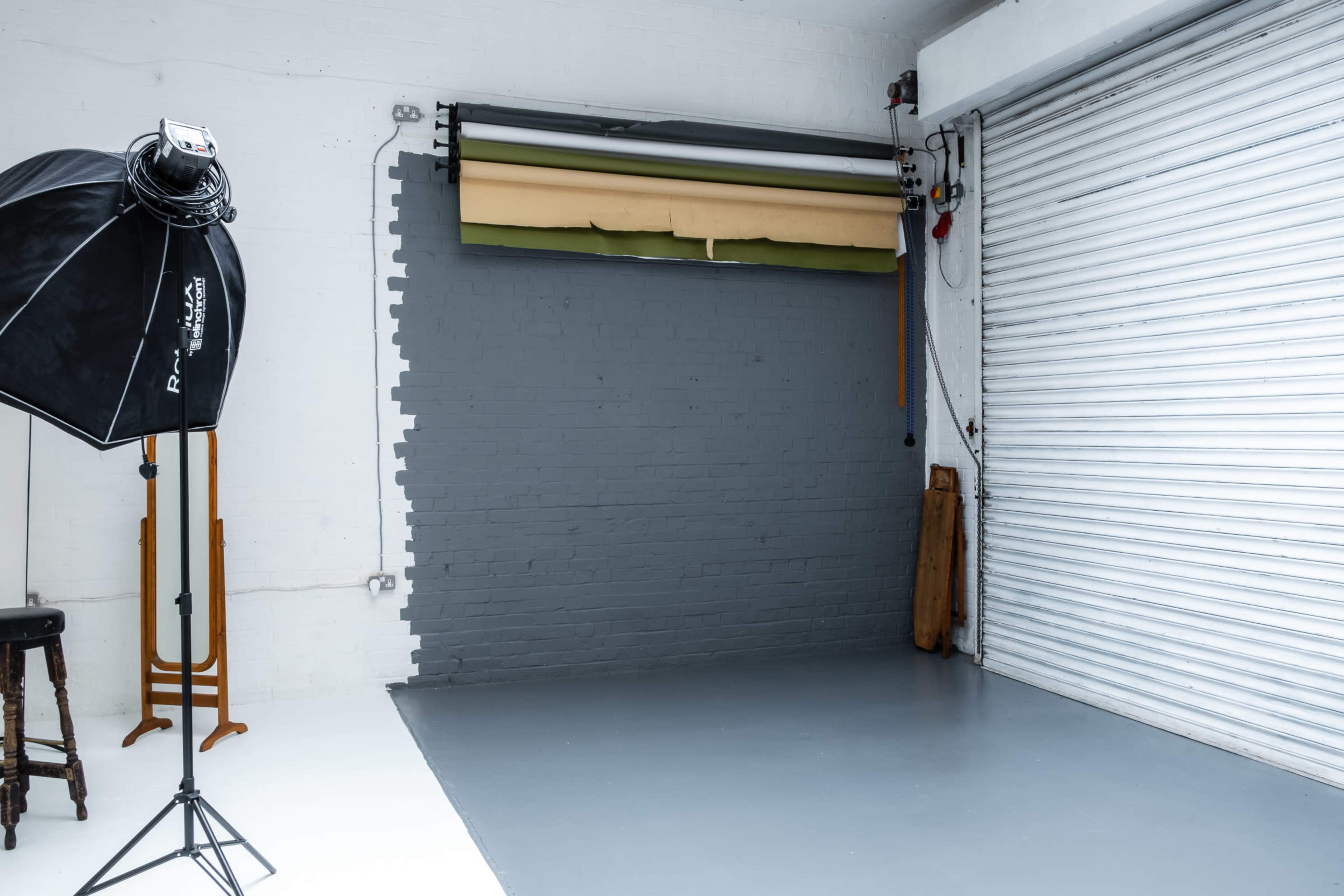A photography studio with a gray wall, a roll-up backdrop rack, and a large softbox light on a stand.