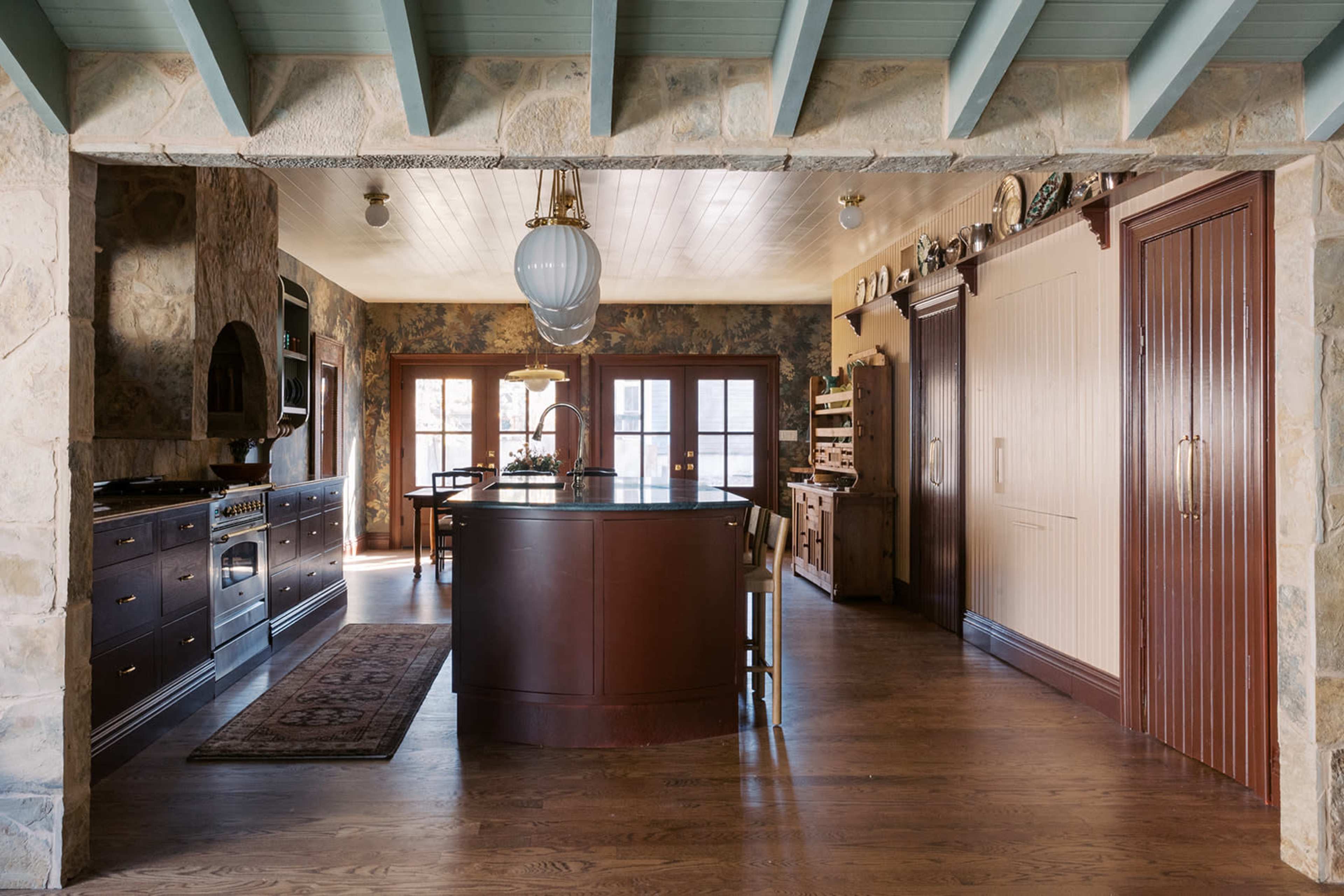 The kitchen features a large dark wooden island, stone walls, and a rustic, open layout with large windows allowing natural light.