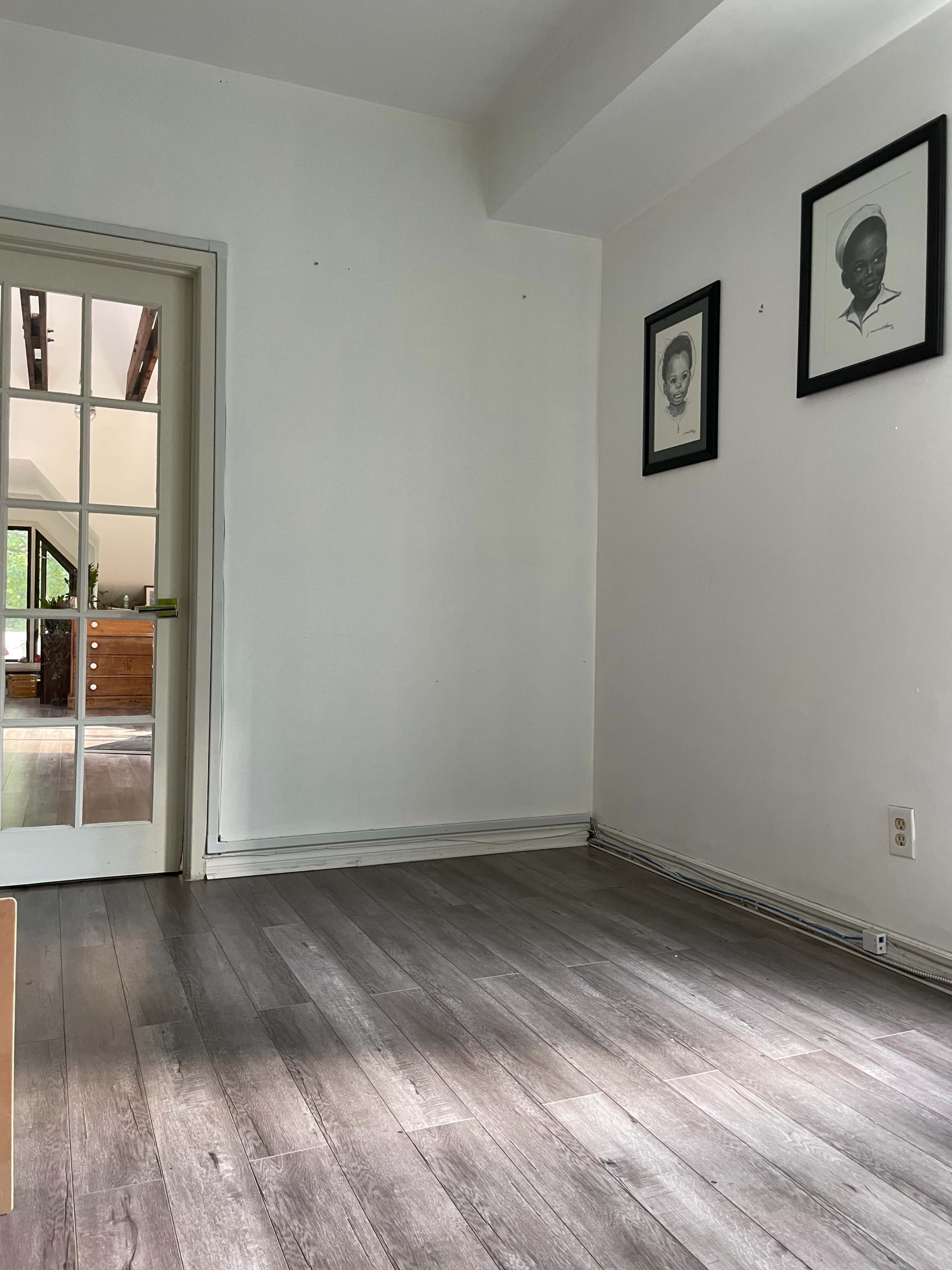 A sparse room with light-colored walls, a wooden floor, and two framed artworks hanging on the wall.