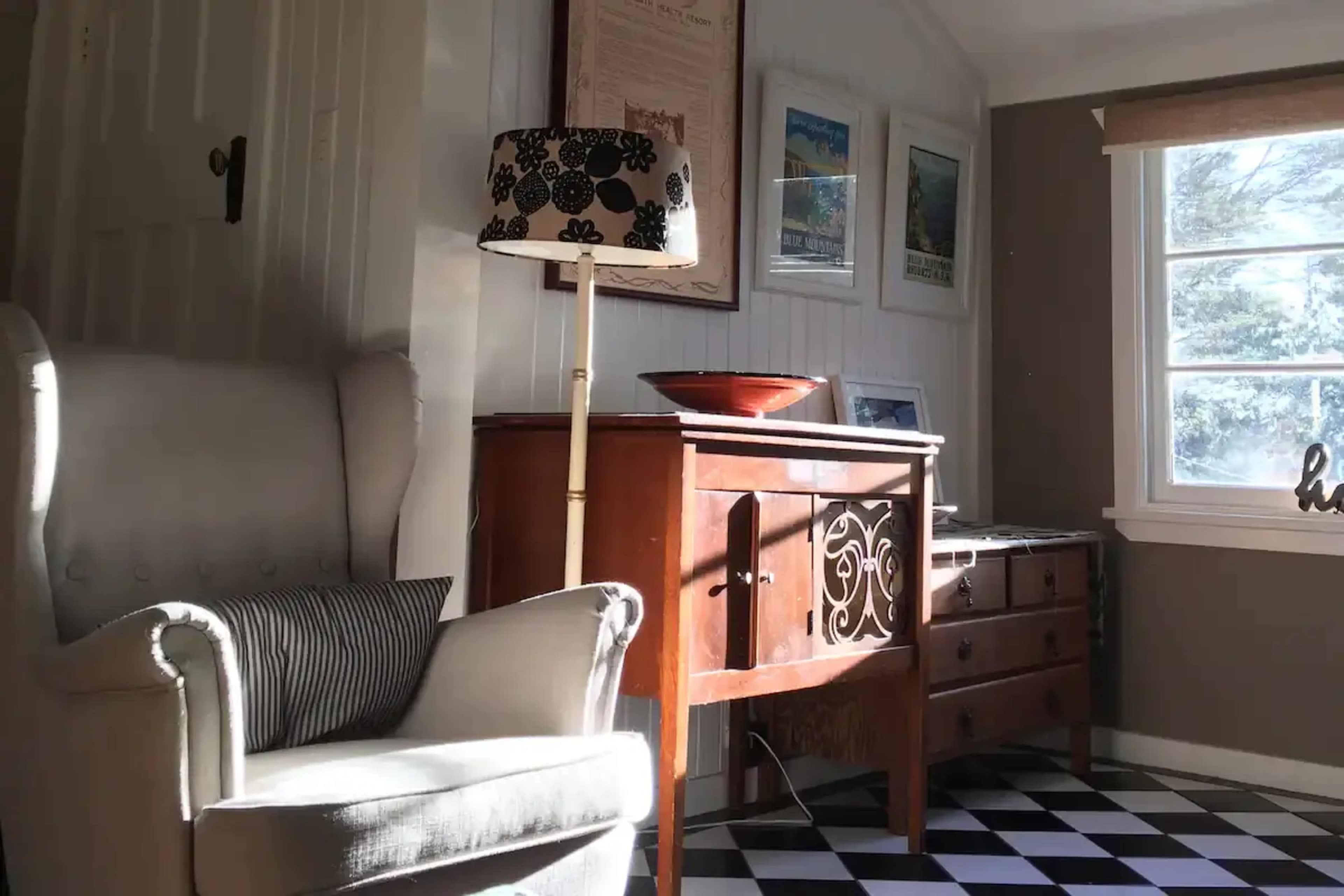 A white armchair sits beside a wooden cabinet and a lamp with a floral shade, against a wall adorned with framed pictures and a window allowing natural light to enter.