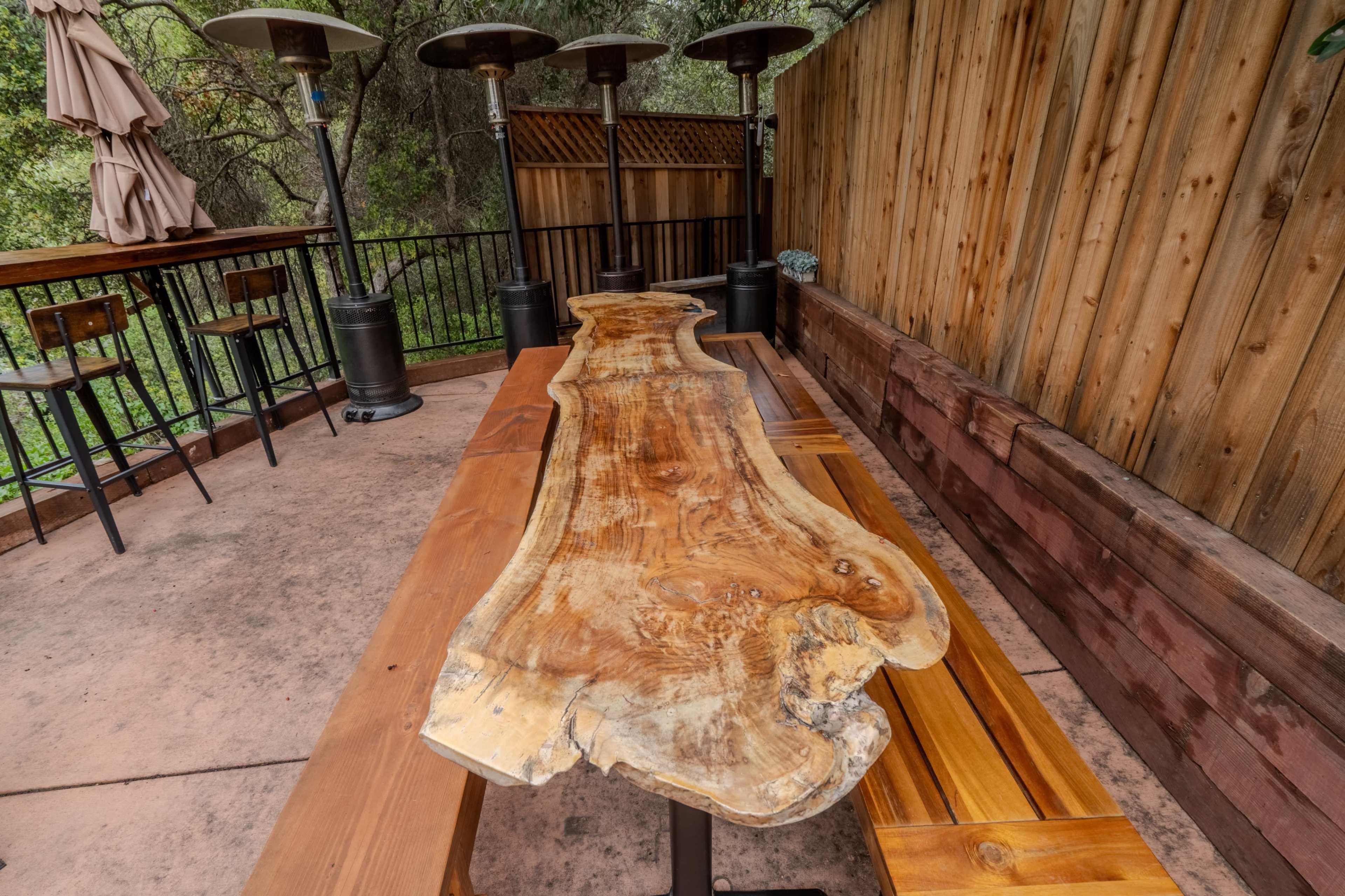 The image shows a long wooden table with a natural edge design set in an outdoor seating area surrounded by wooden walls and patio heaters.