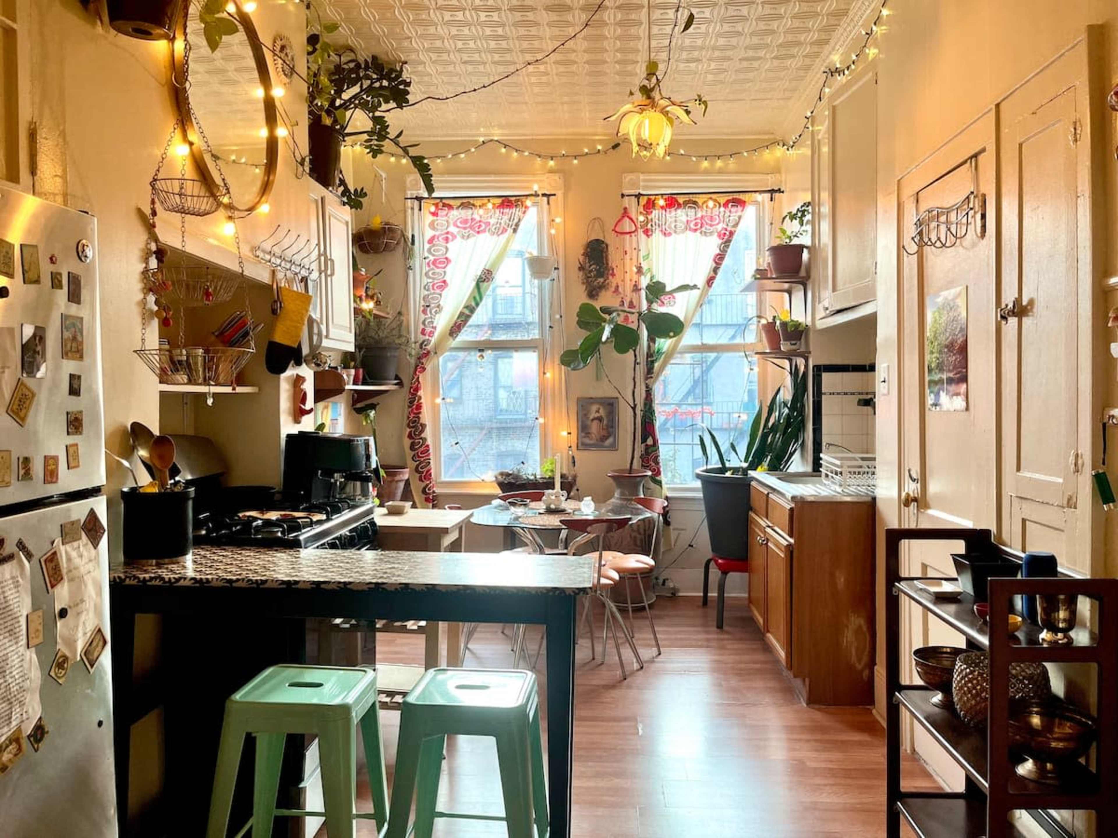 The image shows a cozy kitchen with a mix of modern and vintage elements, featuring a stove, a dining table with chairs, and large windows adorned with plants and curtains.