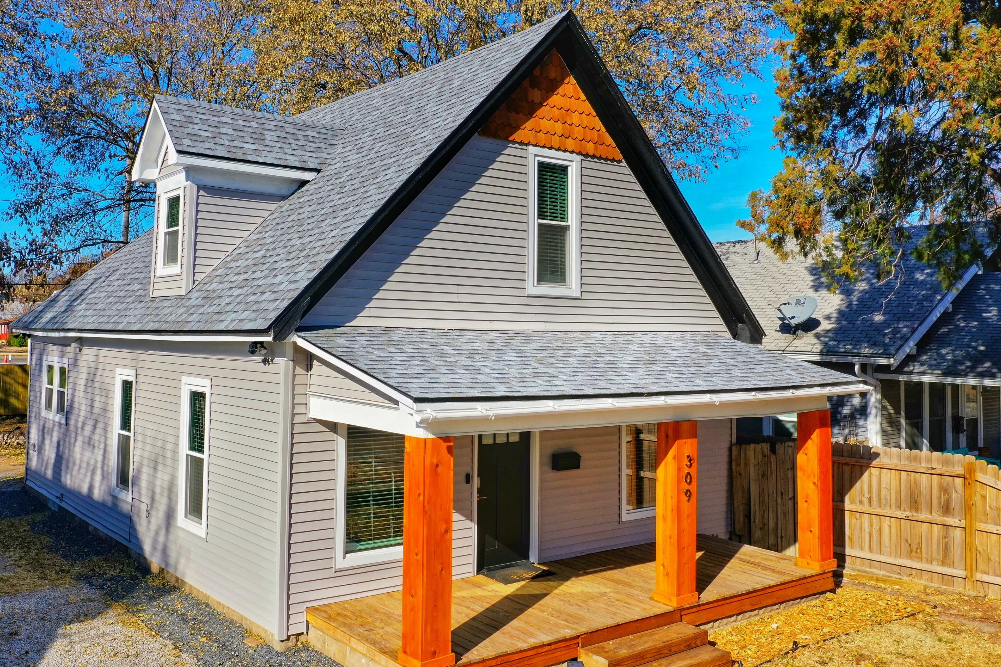 A two-story gray house with a peaked roof and wooden porch is set against a clear blue sky.
