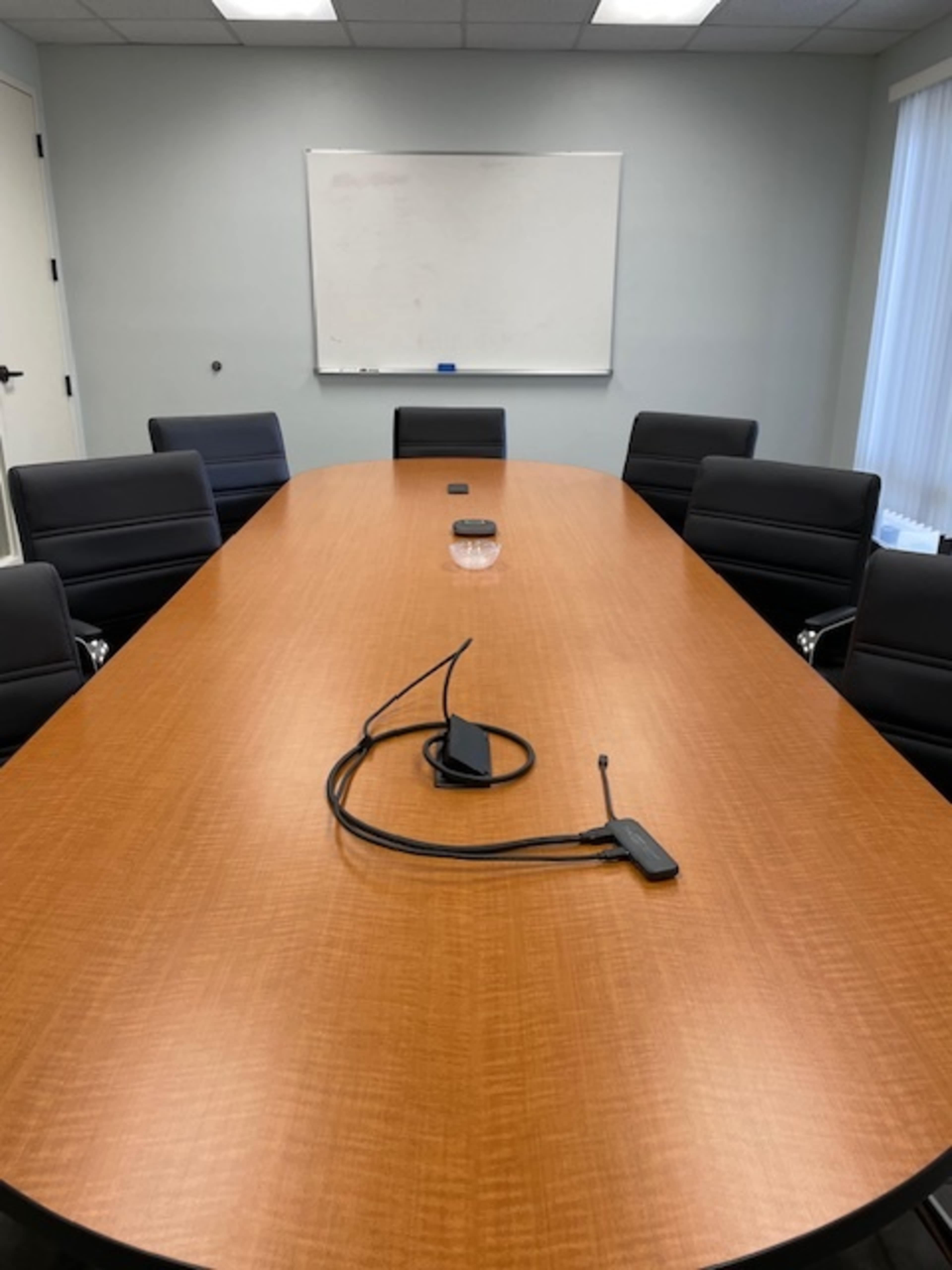 A boardroom features a long wooden table surrounded by black leather chairs, with a whiteboard visible on the wall behind it.