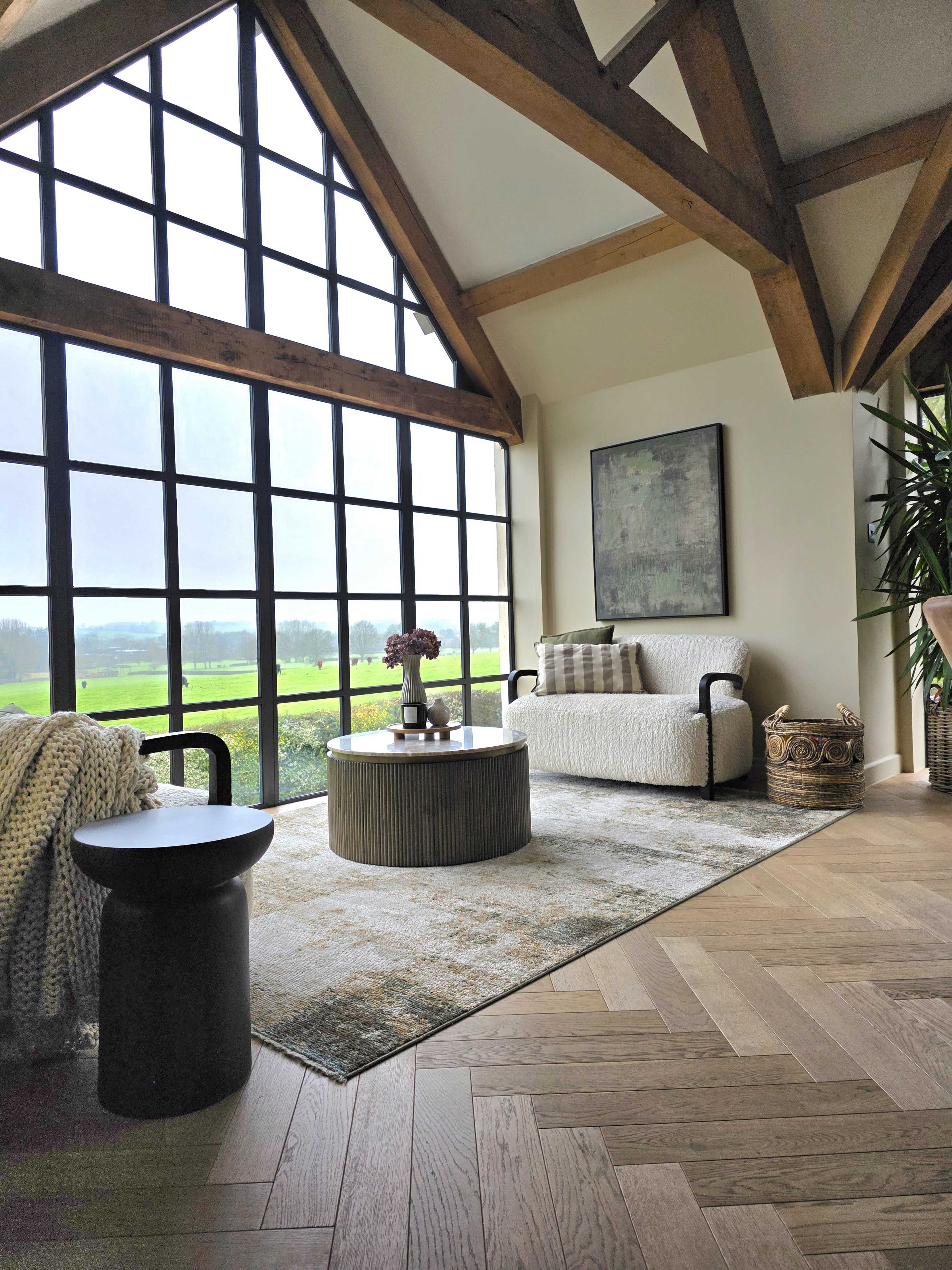 The image shows a spacious living area with large windows, a round coffee table, a sofa, and a view of a green landscape outside.