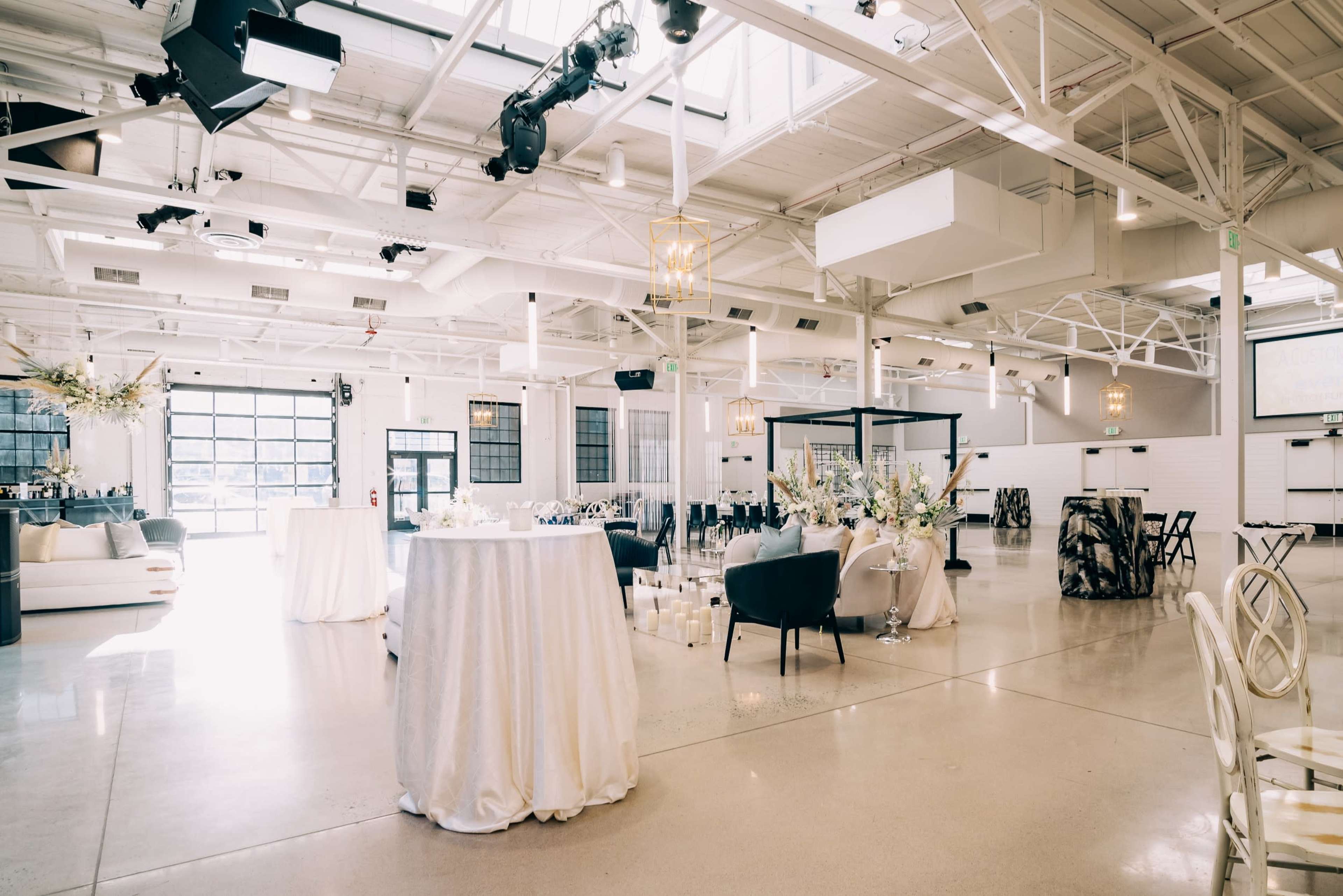 A spacious event venue featuring elegantly arranged tables, chairs, and decorative elements under a bright, airy atmosphere created by large windows.