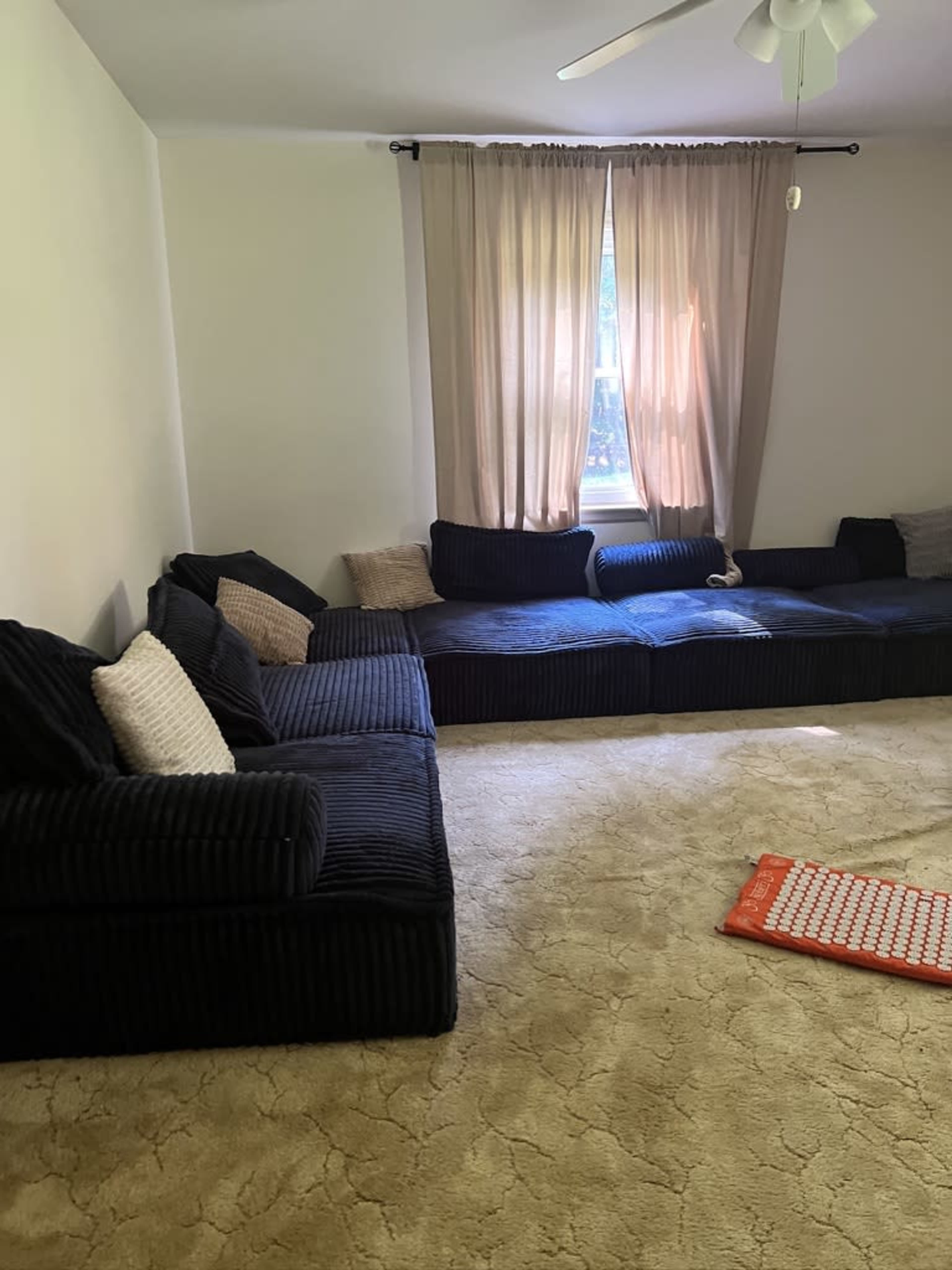 A spacious living room features a dark sectional couch and large window with sheer curtains, illuminating the beige carpet.
