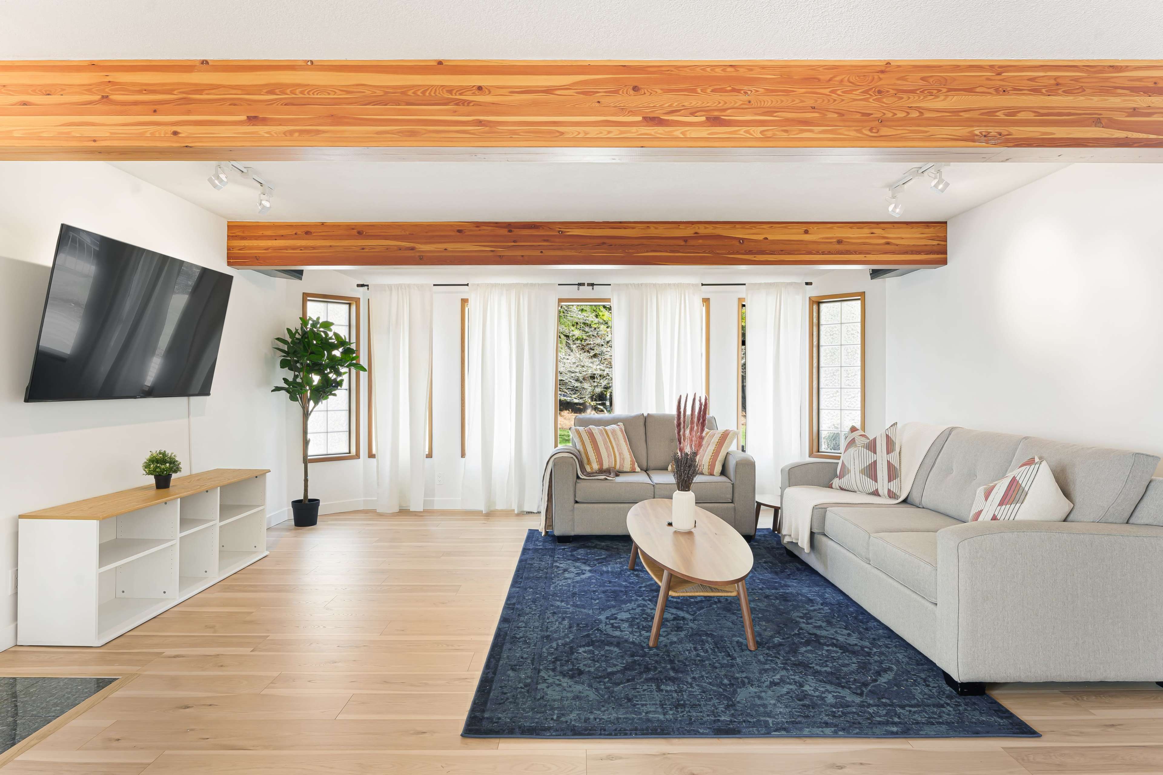 The room features a modern living area with a large television mounted on the wall, a sectional sofa, a coffee table, and large windows allowing natural light to flow in.
