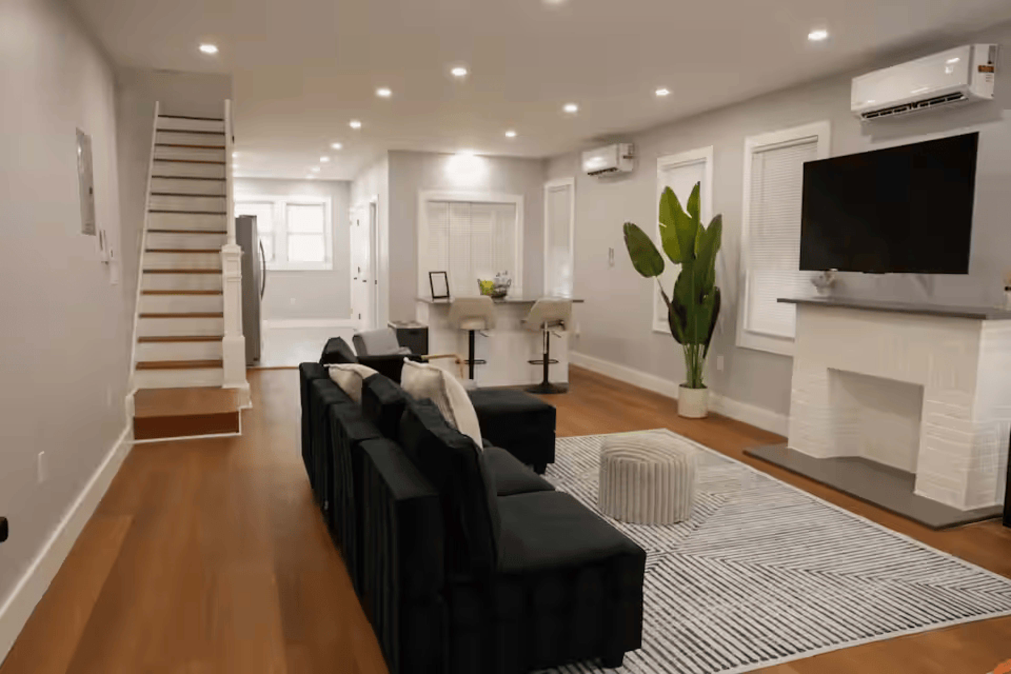 The image shows a modern living room with a black couch, a white fireplace, a large television mounted on the wall, a striped rug on wooden flooring, and a staircase in the background.