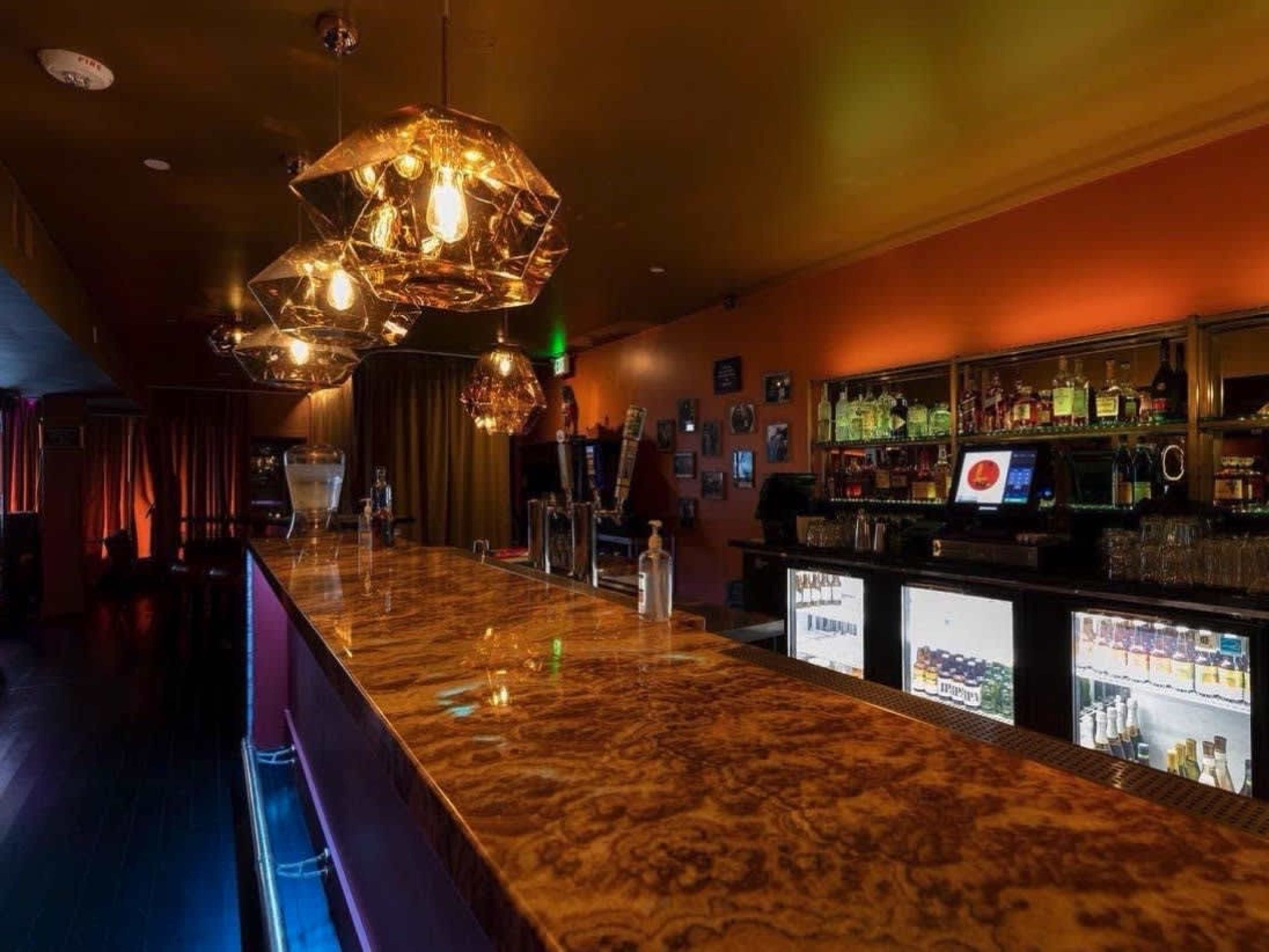 The image shows a bar area with a polished countertop, illuminated pendant lights, and shelves lined with bottles in a dimly lit setting.