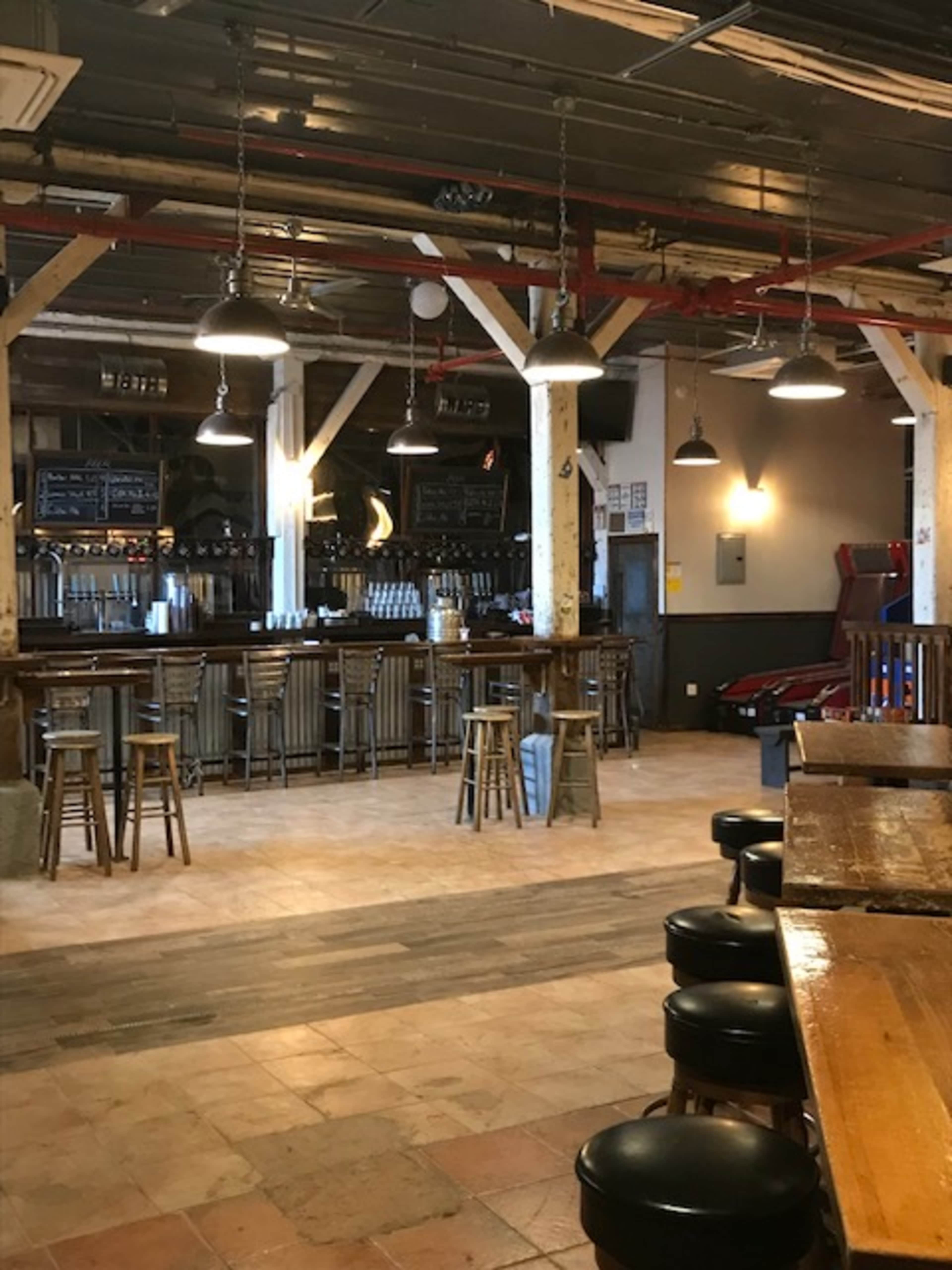 A rustic bar interior with wooden beams, metal stools, and a spacious layout featuring a long bar counter and arcade games in the background.
