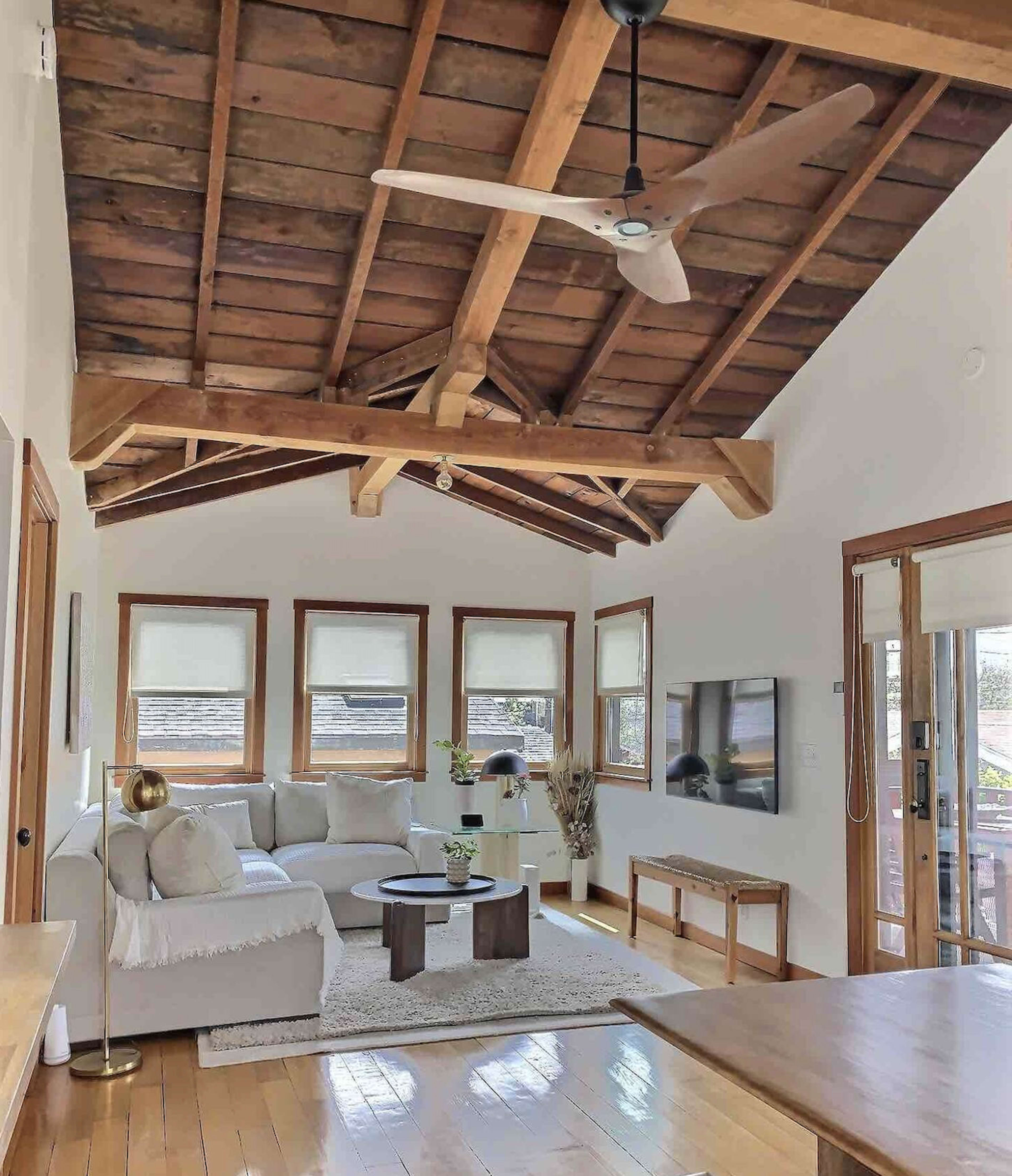 A bright living room with a vaulted wooden ceiling, a light-colored sofa, a coffee table, and a television mounted on the wall.