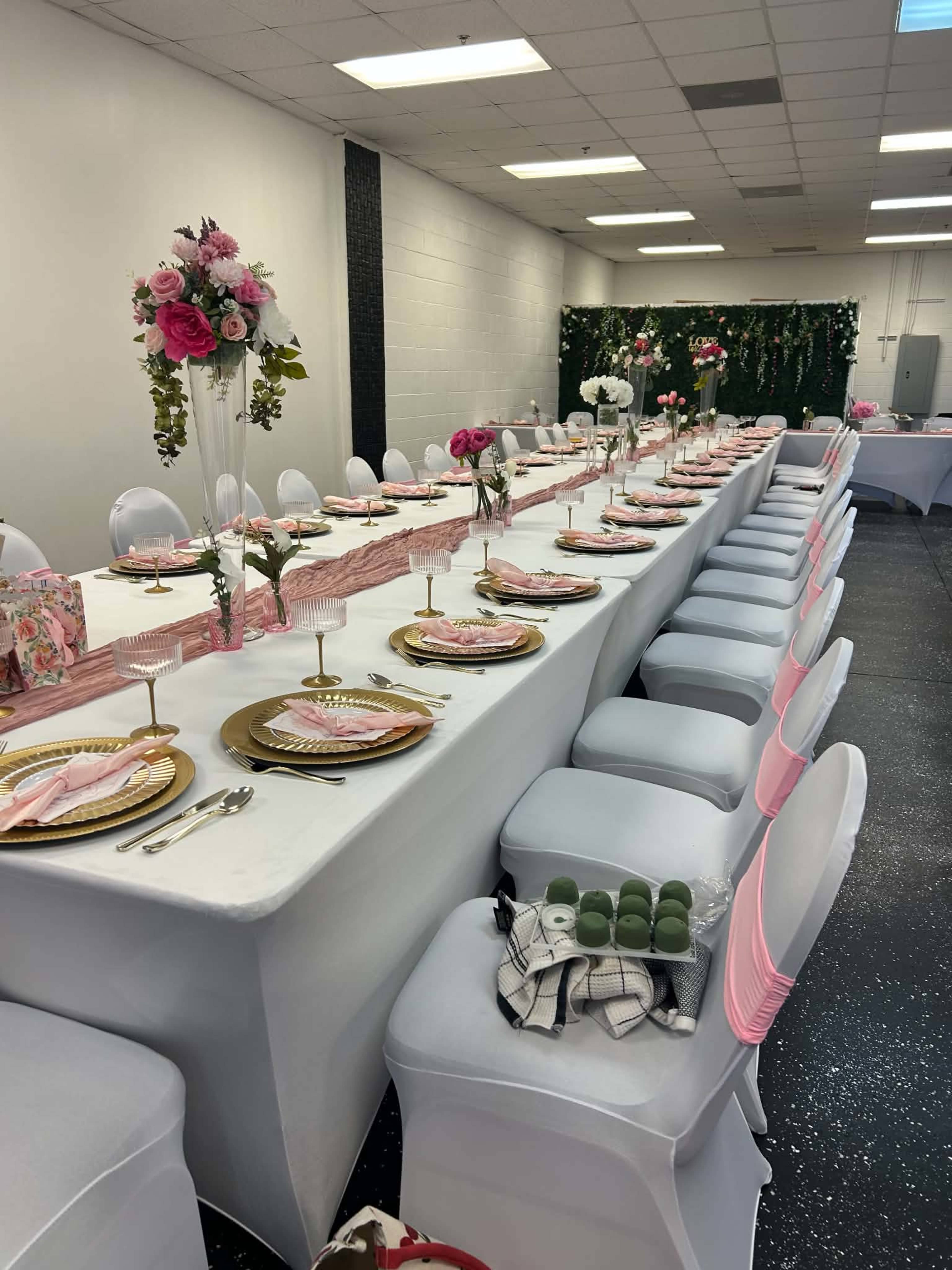 The image shows a long, elegantly set dining table adorned with floral centerpieces, gold plates, and pink napkins in a well-lit room.