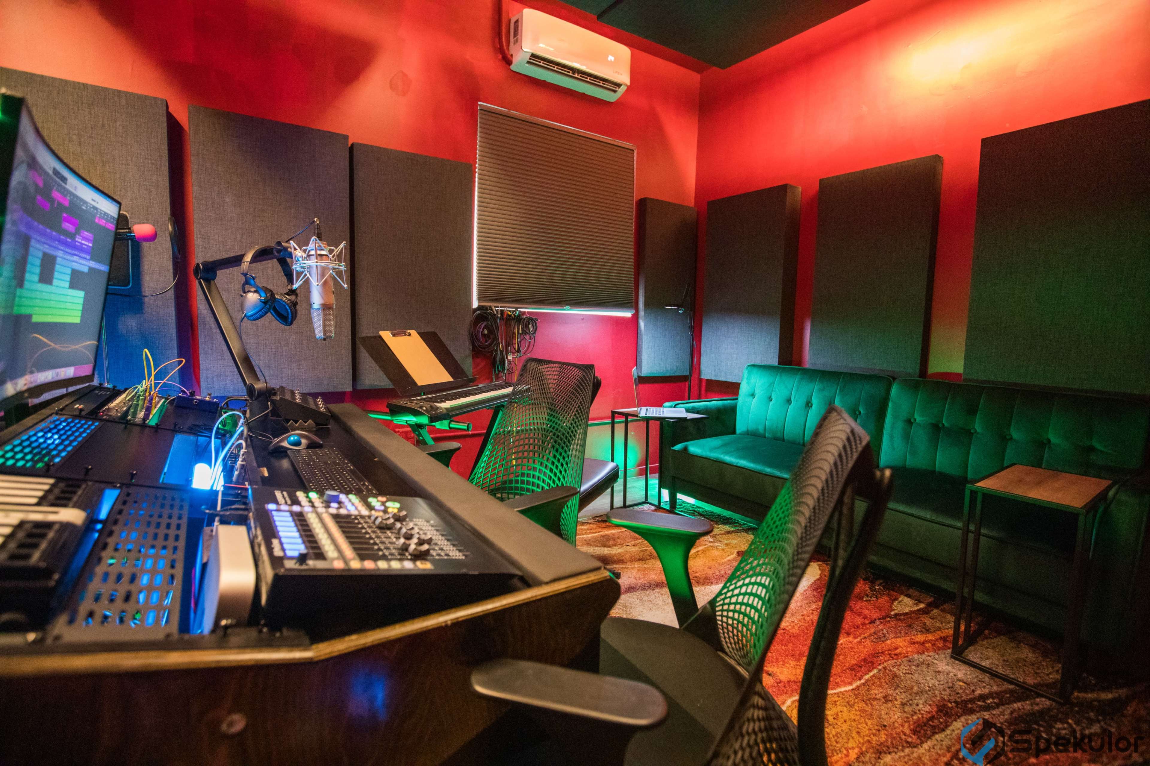 A music studio features a mixing console, microphones, and a green couch against red walls, with acoustic panels and a keyboard setup in the background.