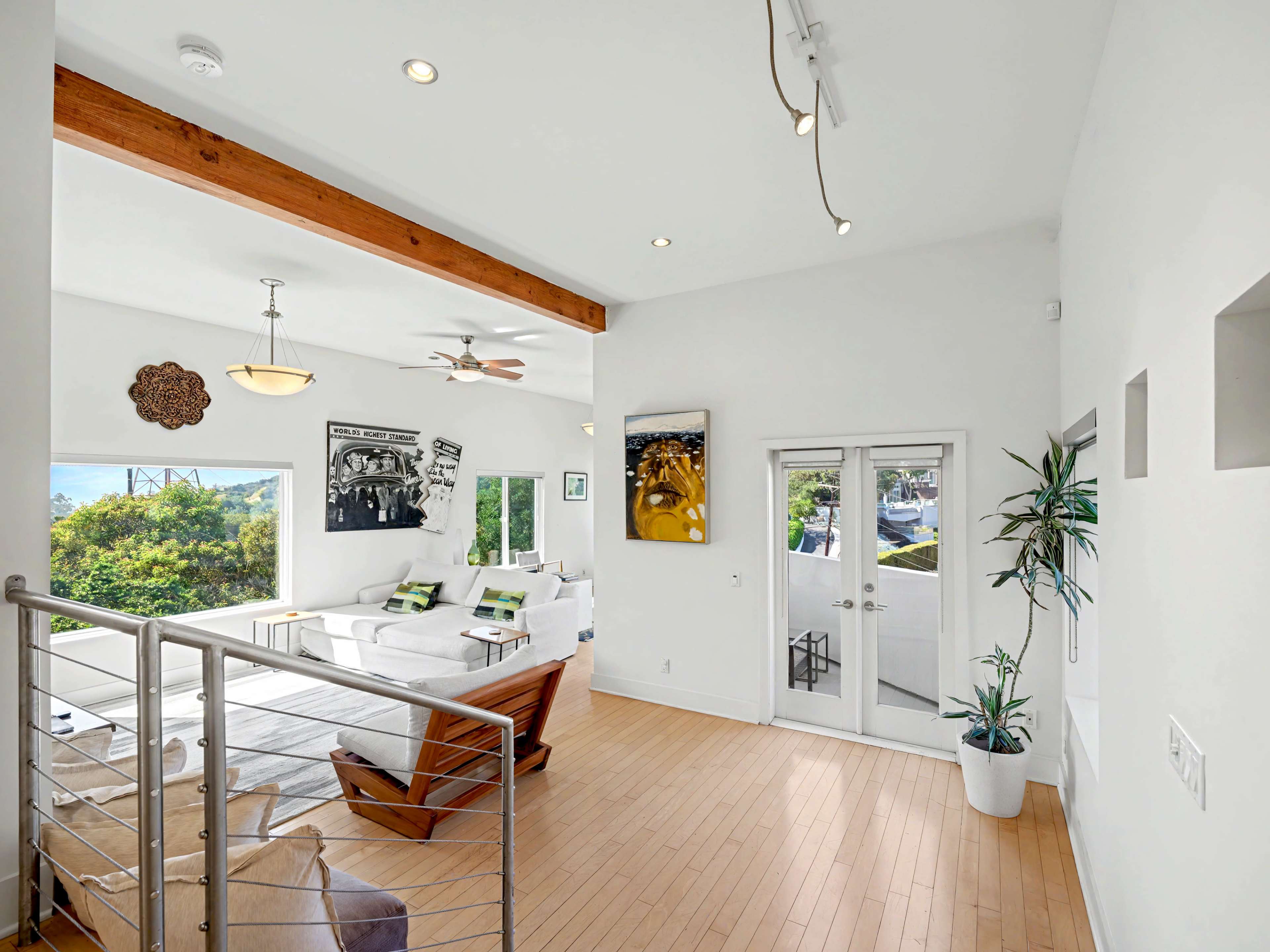 The image shows a bright, modern living room with wooden floors, large windows, and minimalist decor featuring a sectional couch and a potted plant.