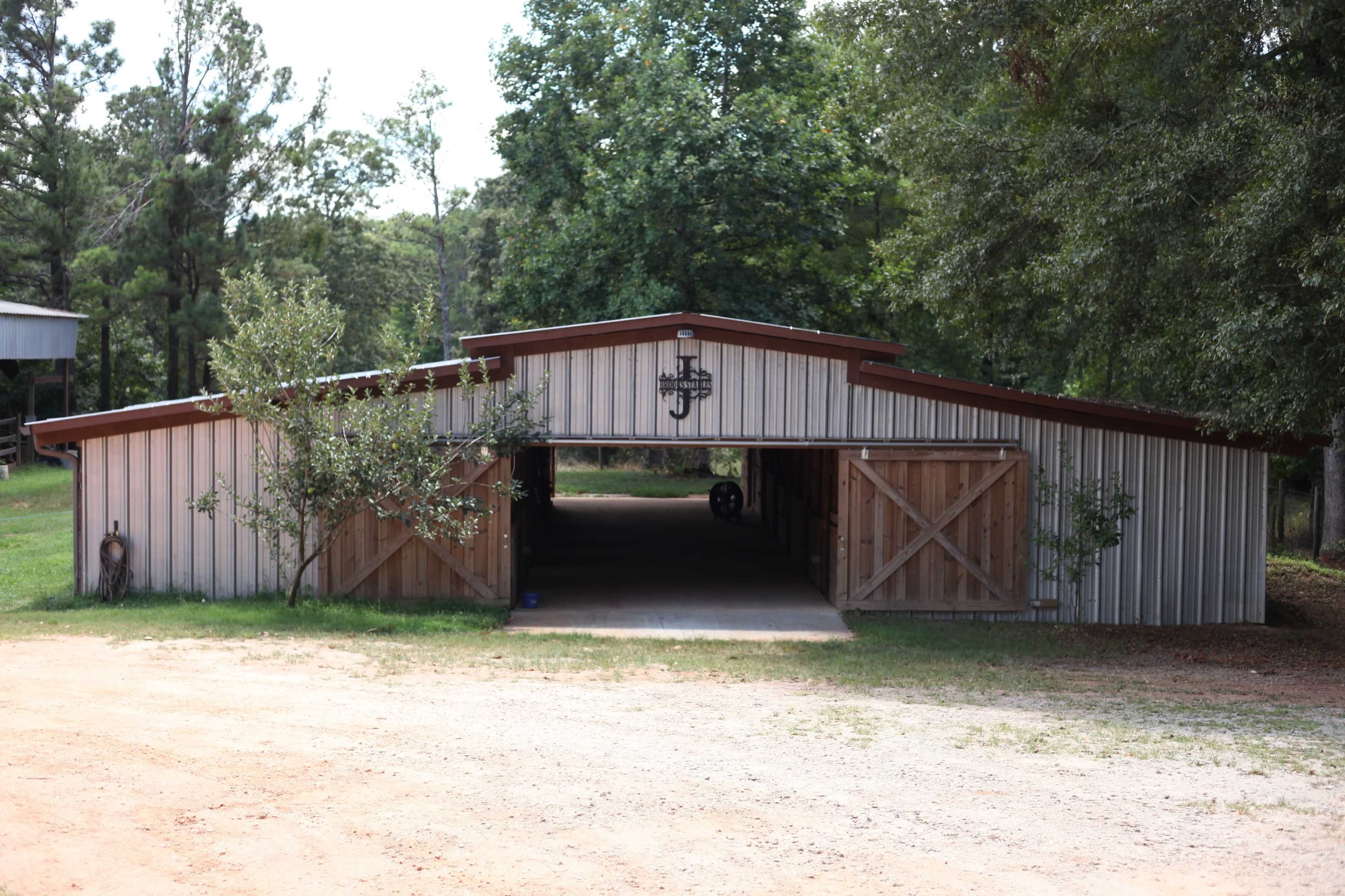 The image shows a large, metal barn with wooden doors and a gravel driveway, surrounded by trees.