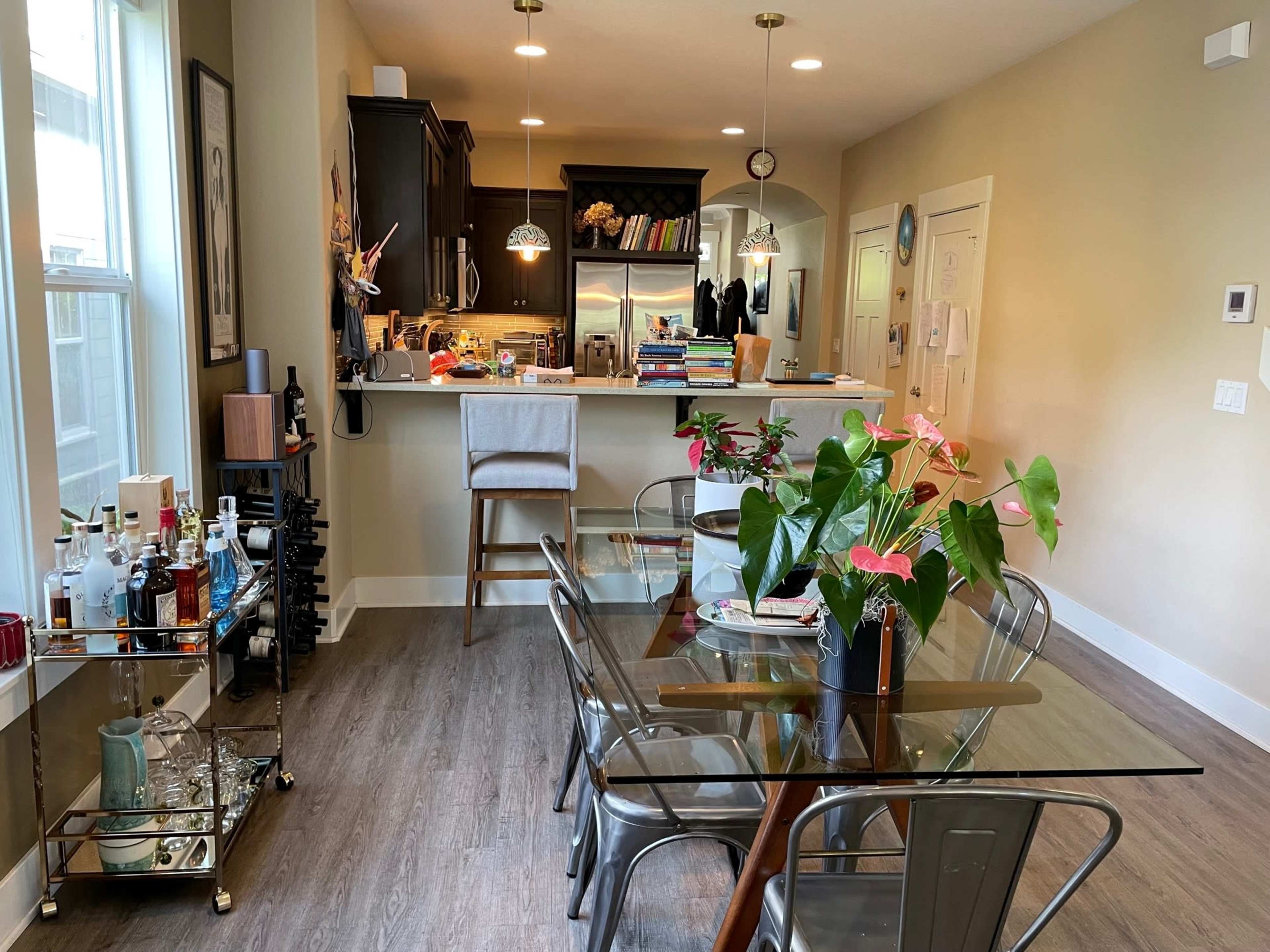 The image shows a modern dining area with a glass-top table surrounded by metal chairs, a bar cart with various bottles, and a kitchen visible in the background.