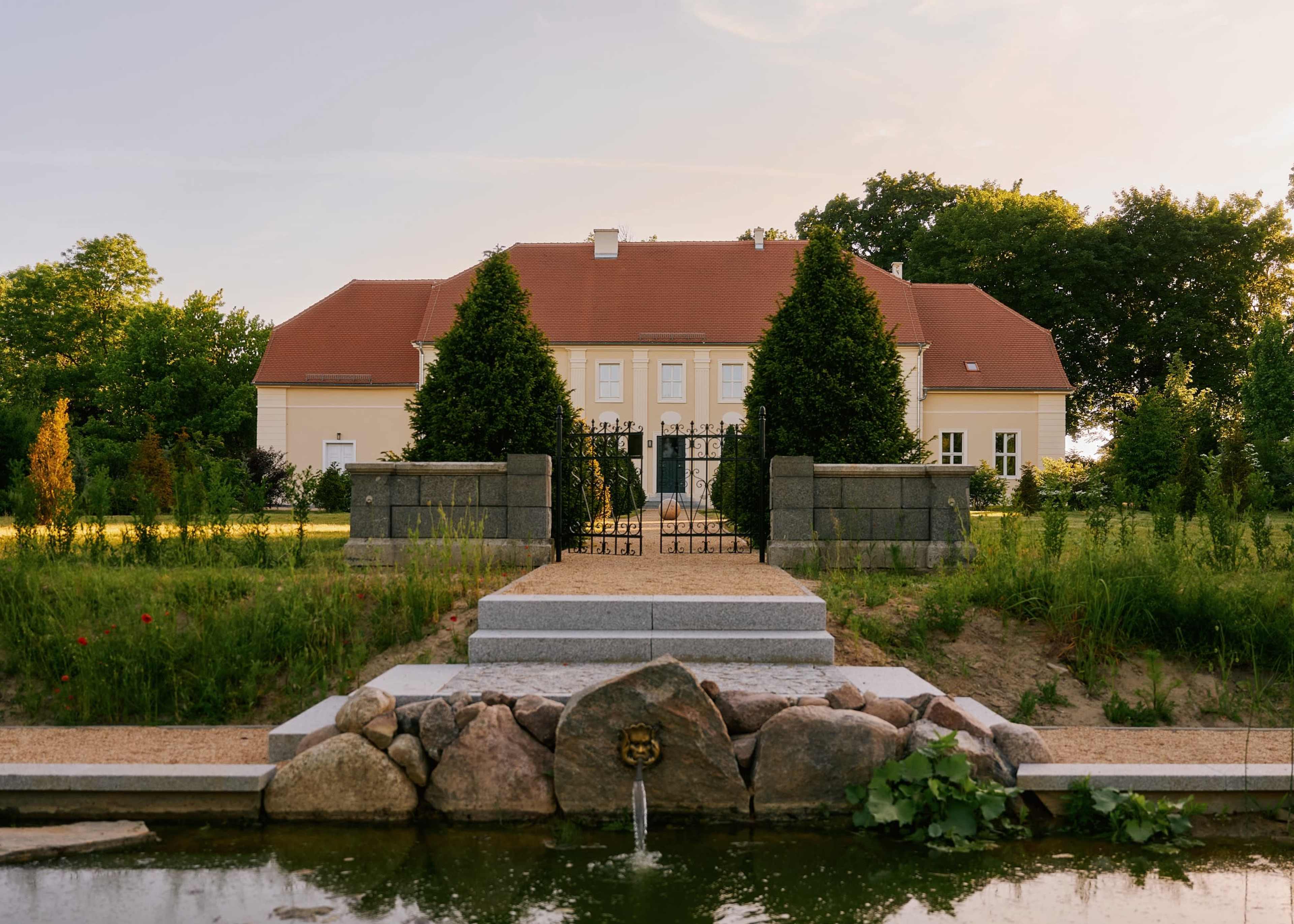A large, two-story building with a red roof is flanked by symmetrical trees and pathways leading to a gated entrance, set in a landscaped garden with a small pond in the foreground.
