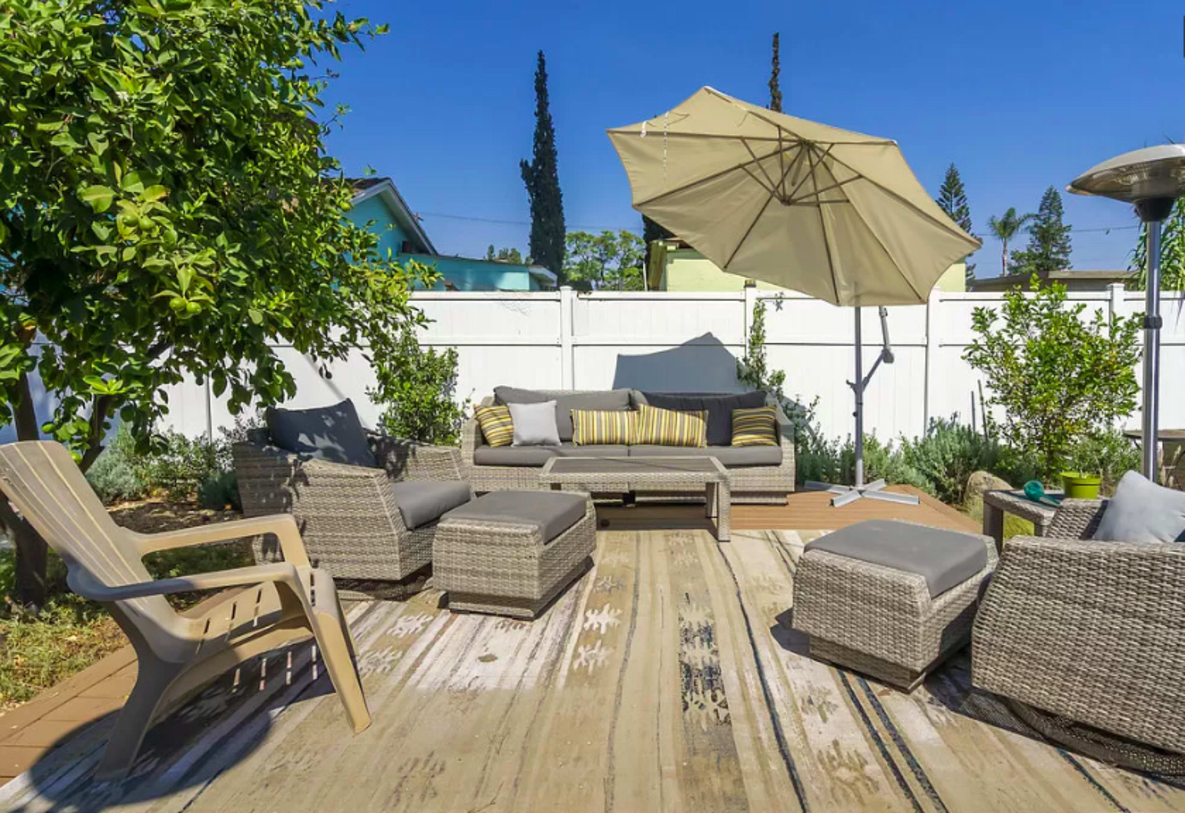 The scene features a patio area with wicker furniture, an umbrella, and a natural rug, surrounded by a white fence and greenery.