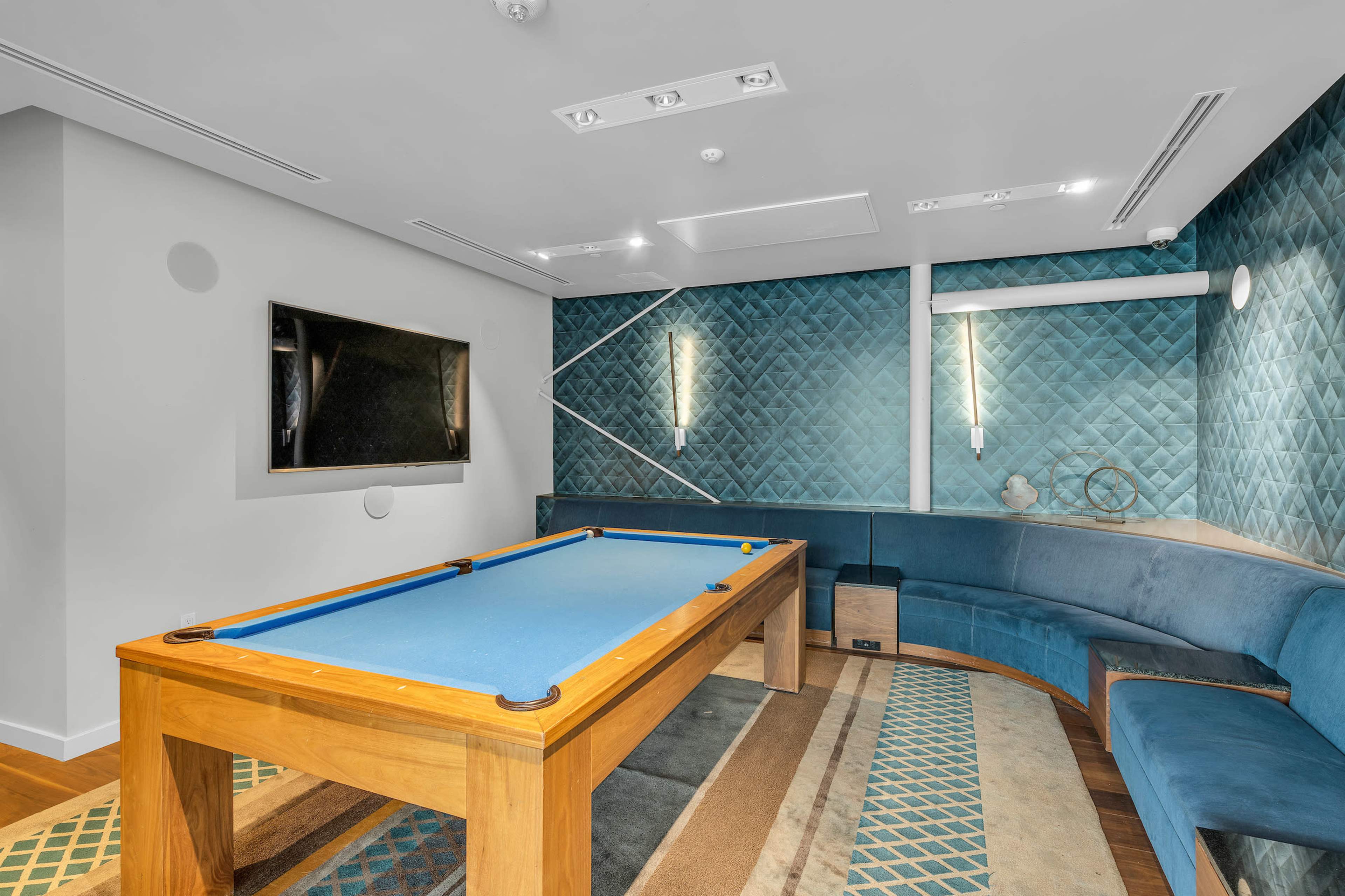 A modern game room featuring a blue pool table, a large wall-mounted television, and a curved seating area against a textured wall.