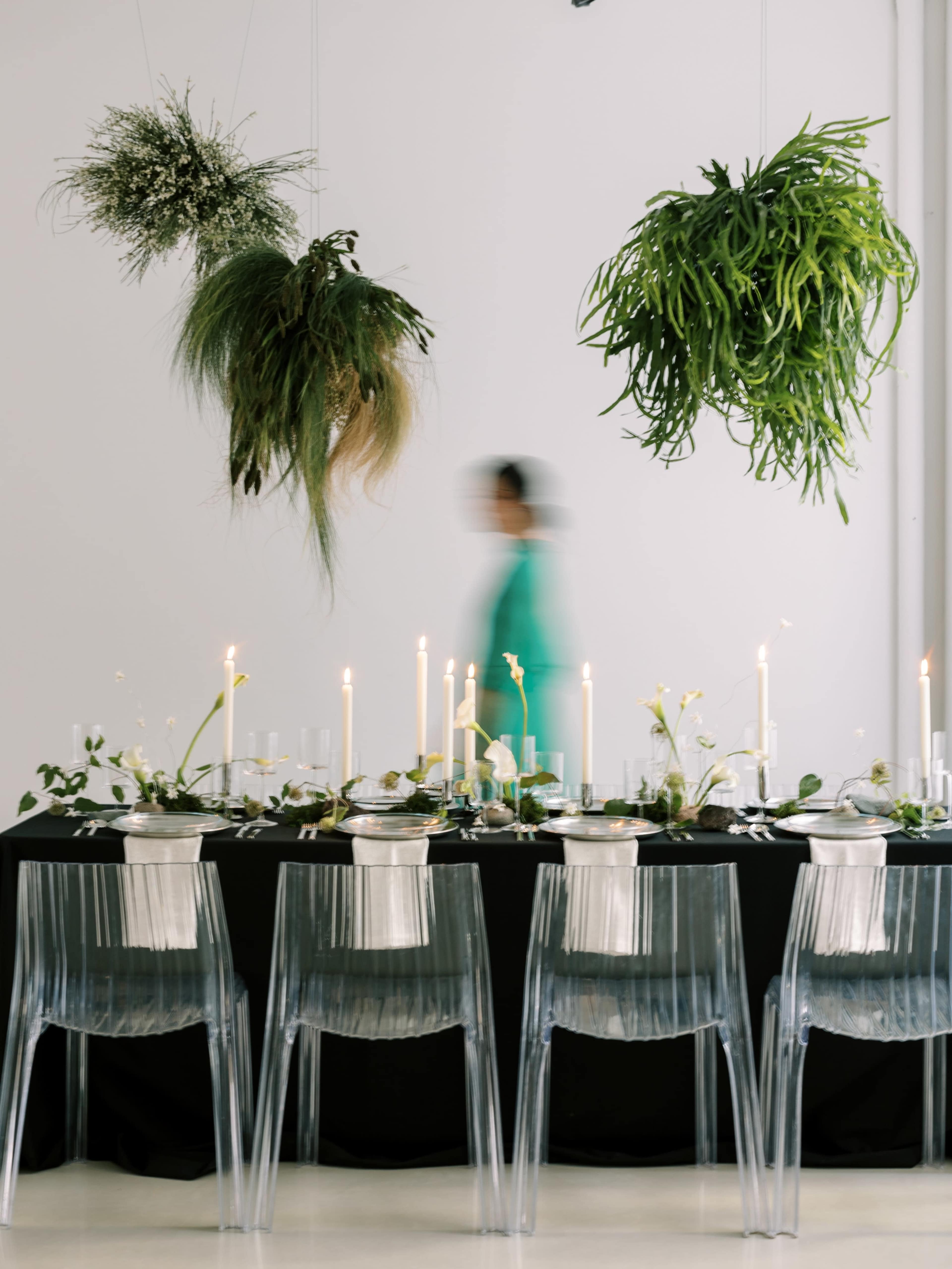 A long dining table set with candles and greenery is partially obscured by a person walking in a teal outfit in a modern space.