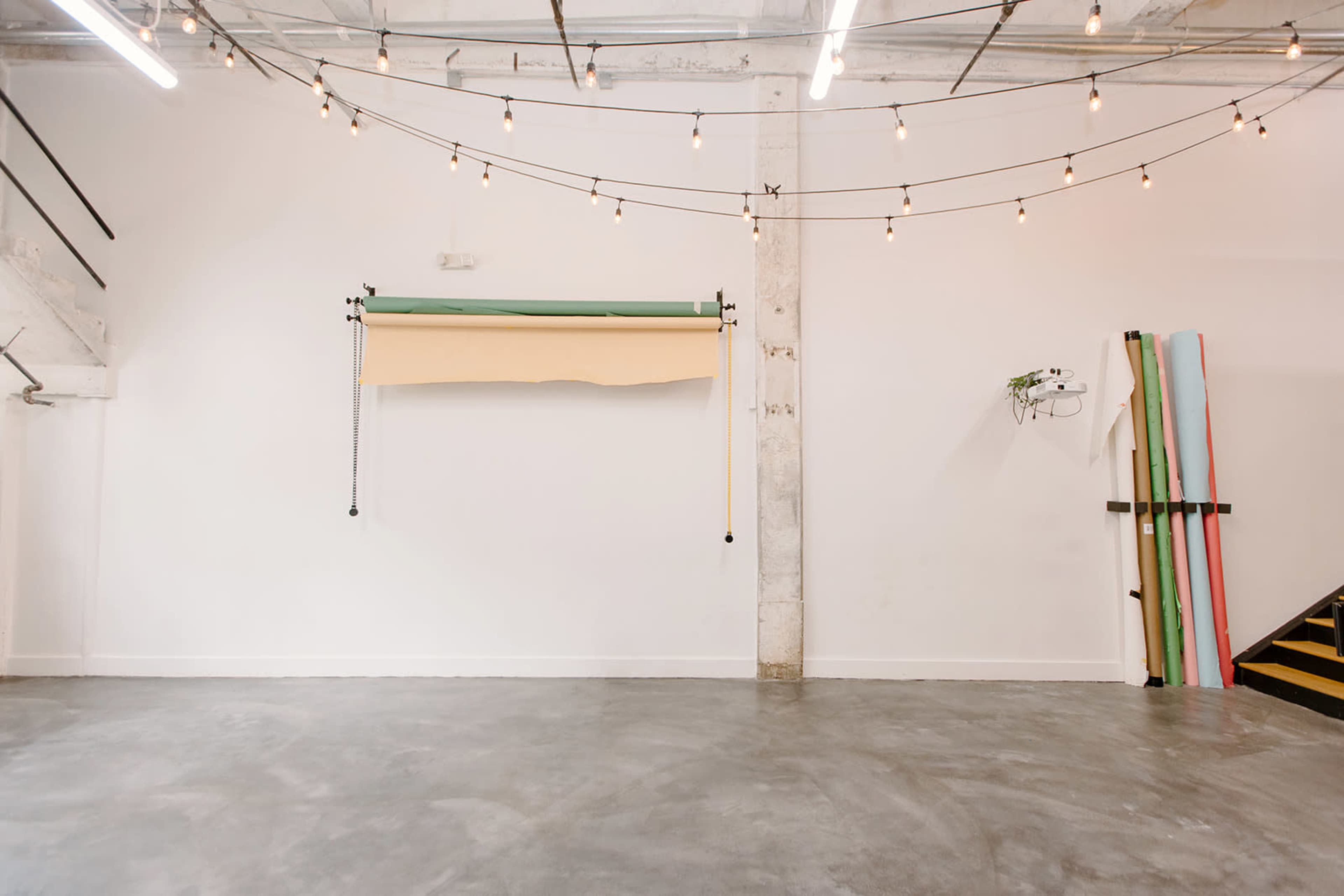 A minimalist studio space with a roll of colored backdrop paper hanging on the wall, string lights overhead, and a clear concrete floor.
